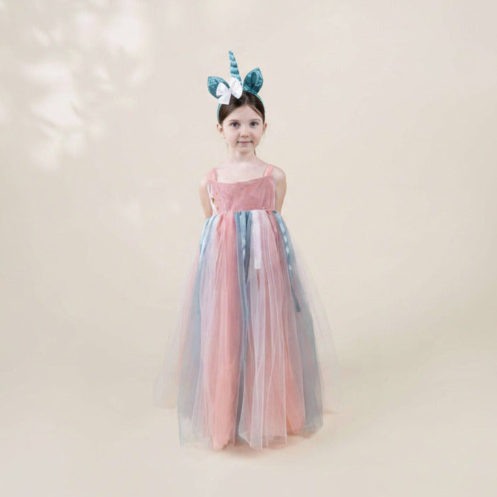 A young girl stands against a soft beige studio background wearing a long tulle dress with pink, blue, and gray panels, plus a matching horn headband.