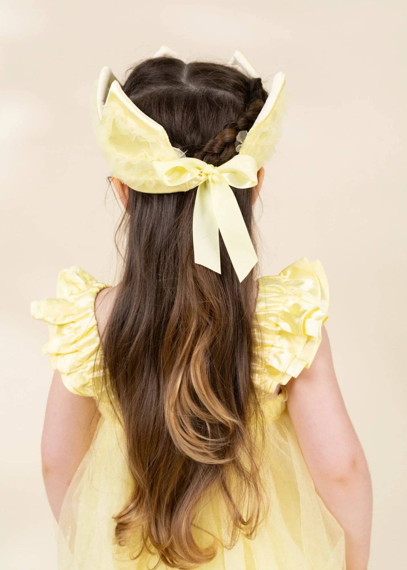 A young girl viewed from behind wearing a pale yellow ruffled bonnet with a large bow and long wavy brown hair, dressed in a matching pale yellow ruffled dress against a soft beige background.