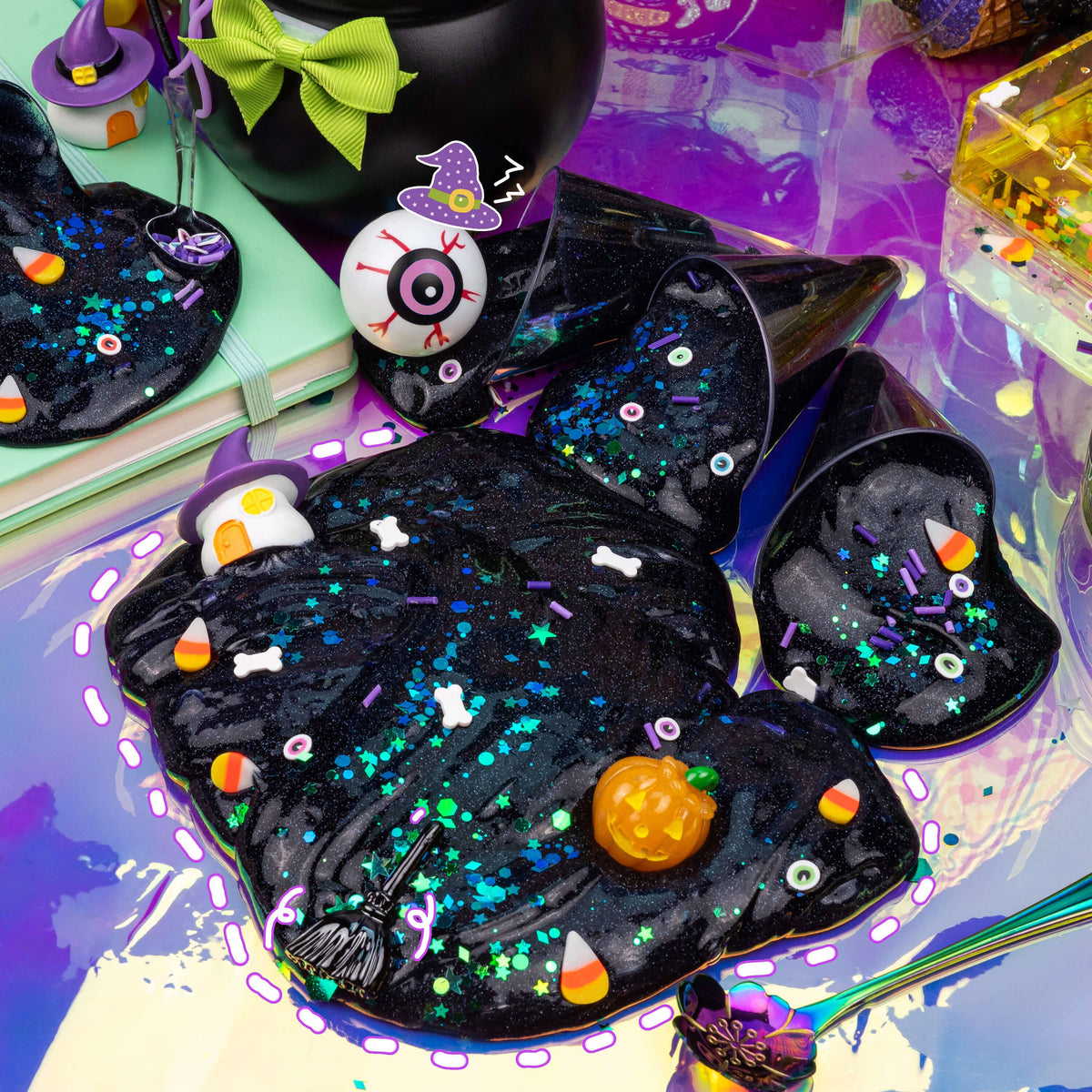 Halloween-themed black slime with glitter, candy corn, bones, and a pumpkin, displayed with matching black party hats and witch accessories on a colorful purple background.