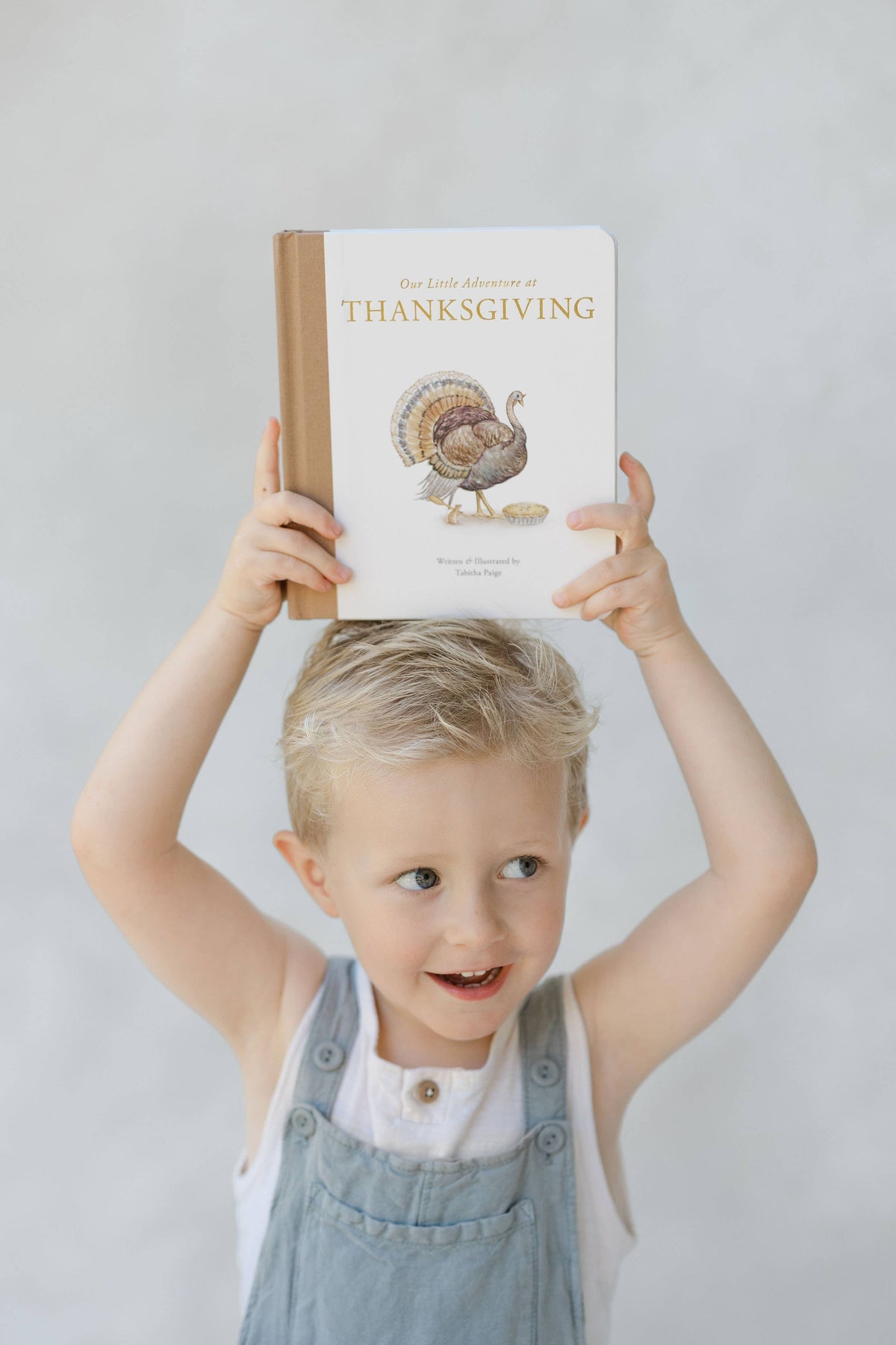 A young blonde child in blue denim overalls holds a hardcover book titled 'The Great Thanksgiving' above their head. The book cover is white with gold lettering and an illustration of a turkey.