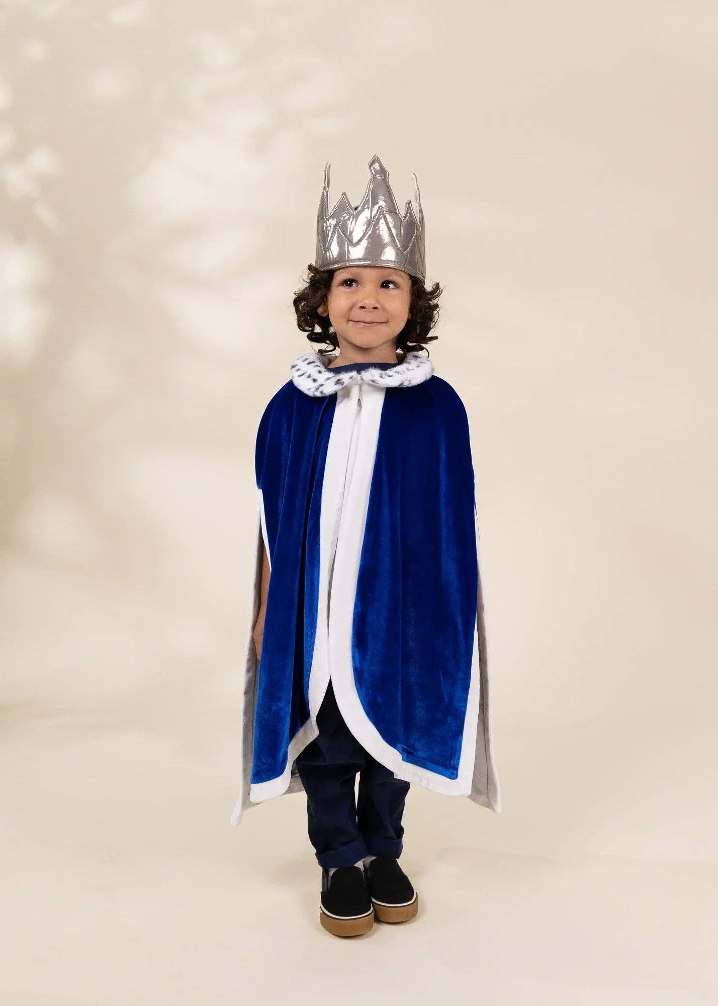 A young child wearing a royal prince costume, featuring a royal blue velvet cape with white trim, a matching silver crown, and a white faux-fur collar, standing against a warm beige studio backdrop.