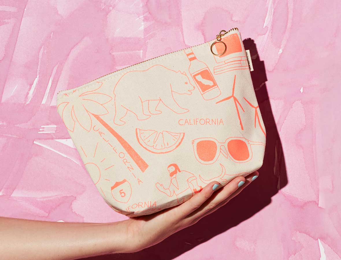 A hand holds a cream-colored zippered pouch decorated with orange line drawings of California-themed motifs, including a bear, palm tree, sunglasses, and a bottle, set against a pink watercolor background.