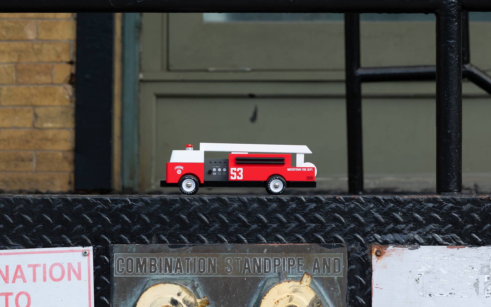 Small red and white model fire truck on a metal surface with a building in the background.