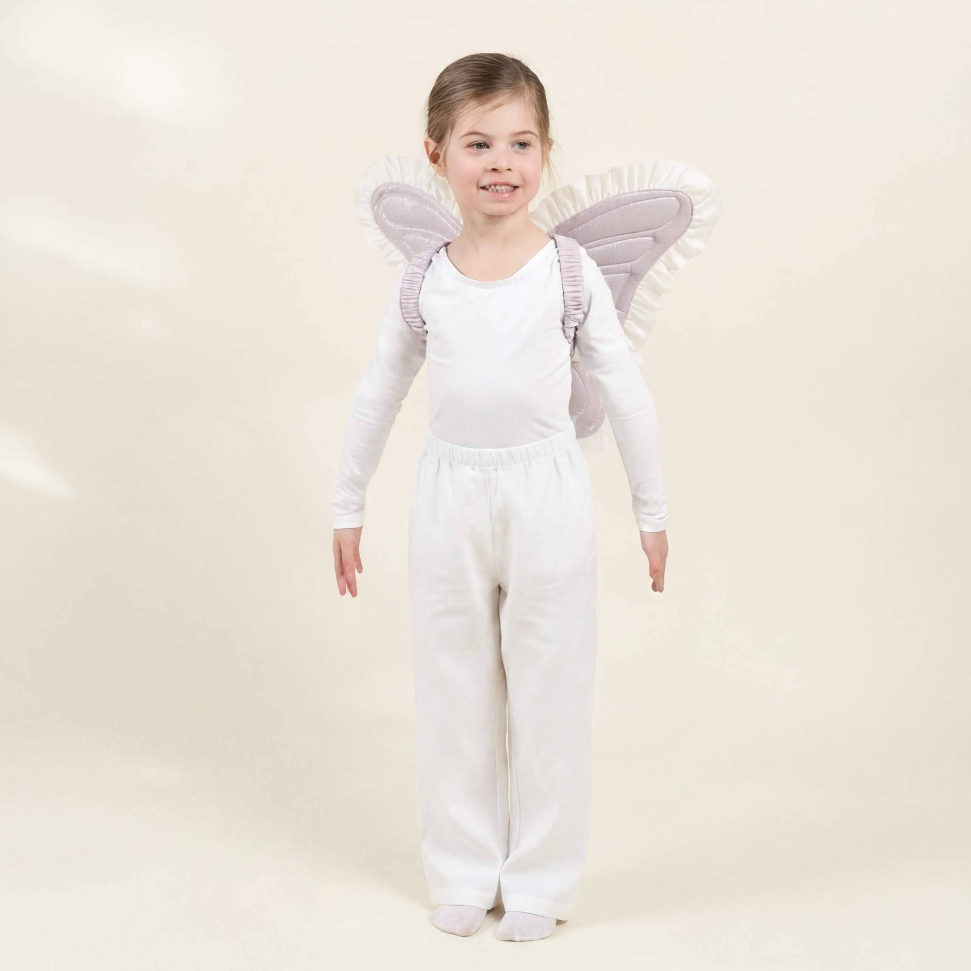A young girl stands against a warm cream studio background wearing a white long-sleeve top and white pants, with soft lavender angel wings attached to her back.