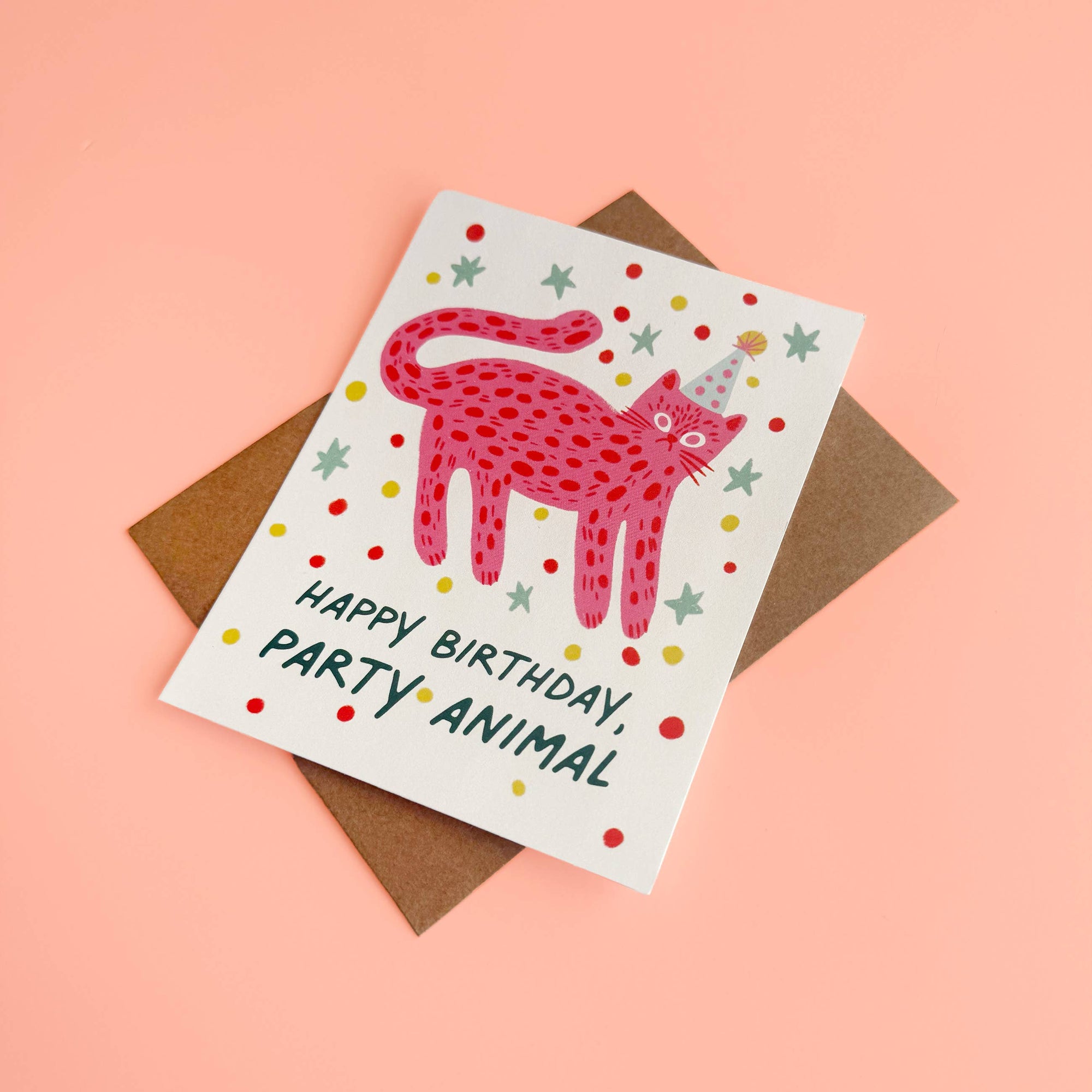 A square birthday card with a white background, featuring a pink spotted cat wearing a party hat and the text 'HAPPY BIRTHDAY, PARTY ANIMAL' in dark green. The card is placed on top of a brown envelope against a solid peach-pink background.
