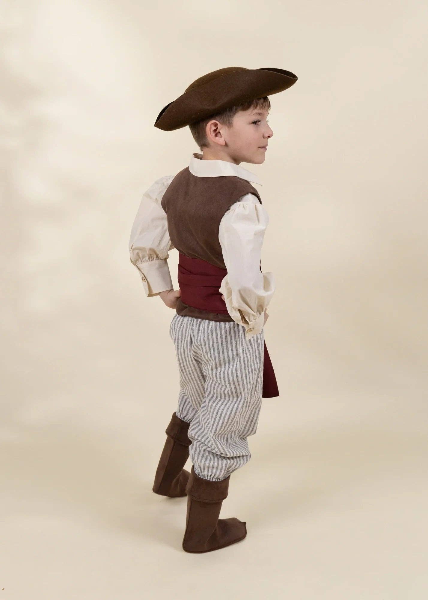 A young boy modeling a pirate costume, shown from a side-back angle against a warm beige studio background. The costume includes a brown tricorne hat, a brown vest over a cream shirt, a dark red waist sash, vertically striped blue and white pants, and brown boot covers.