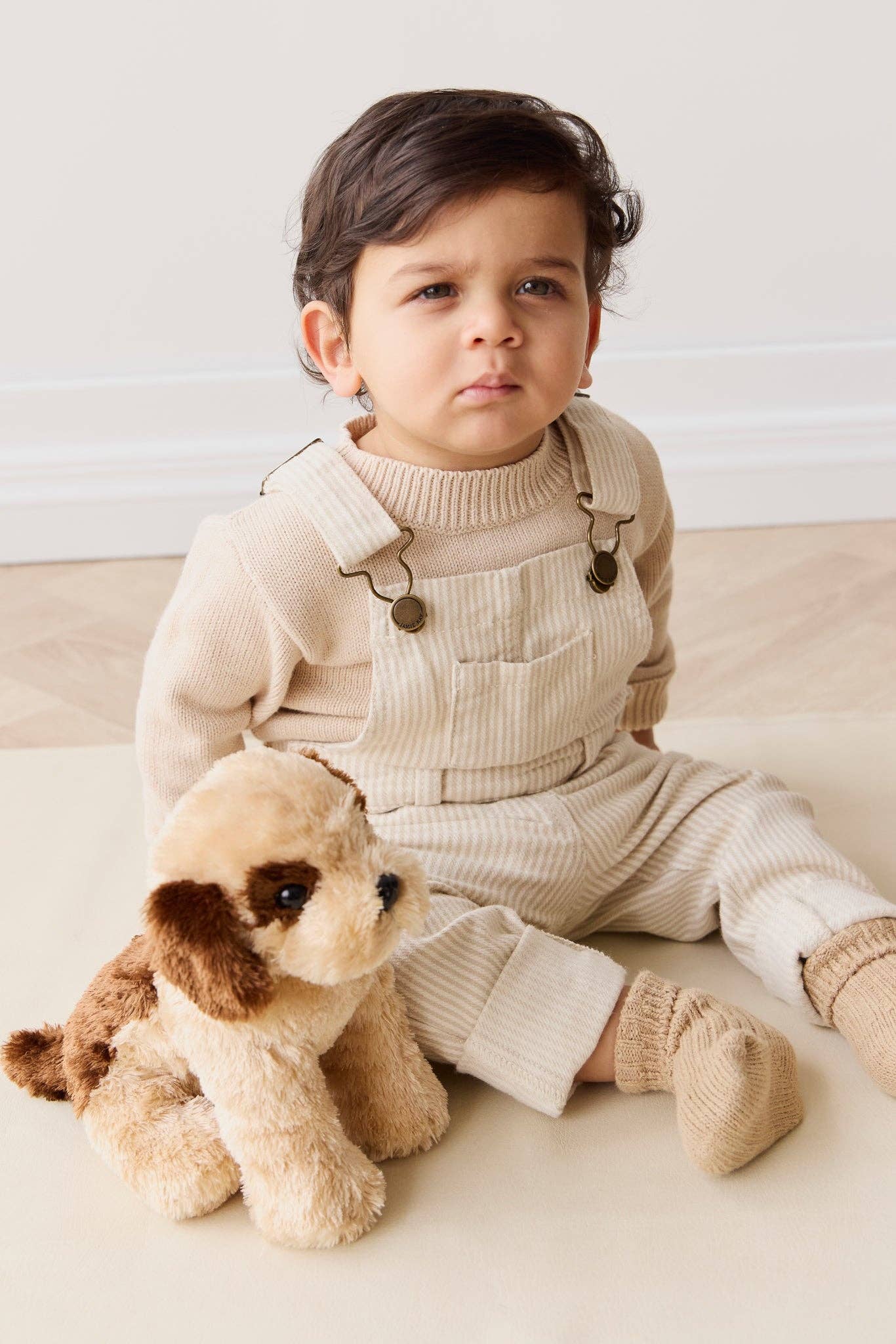 Child wearing a beige outfit with suspenders sitting next to a plush toy on a light background