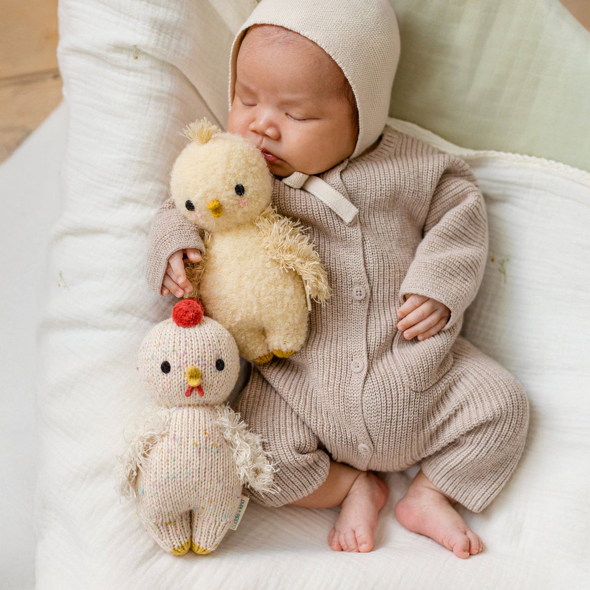 Baby in a knitted outfit holding two plush toys on a soft surface