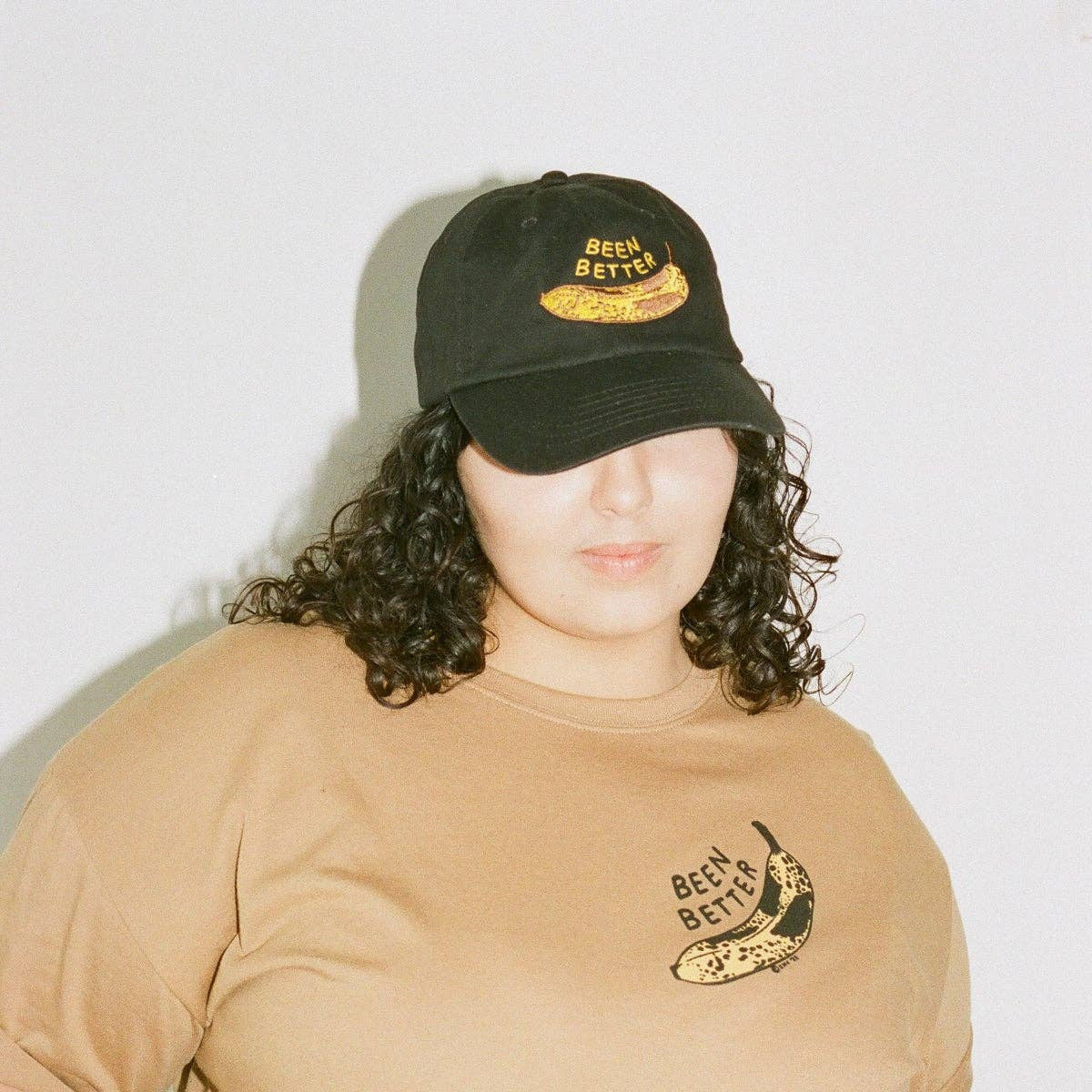 Person wearing a beige t-shirt with a logo and a black cap on a plain background