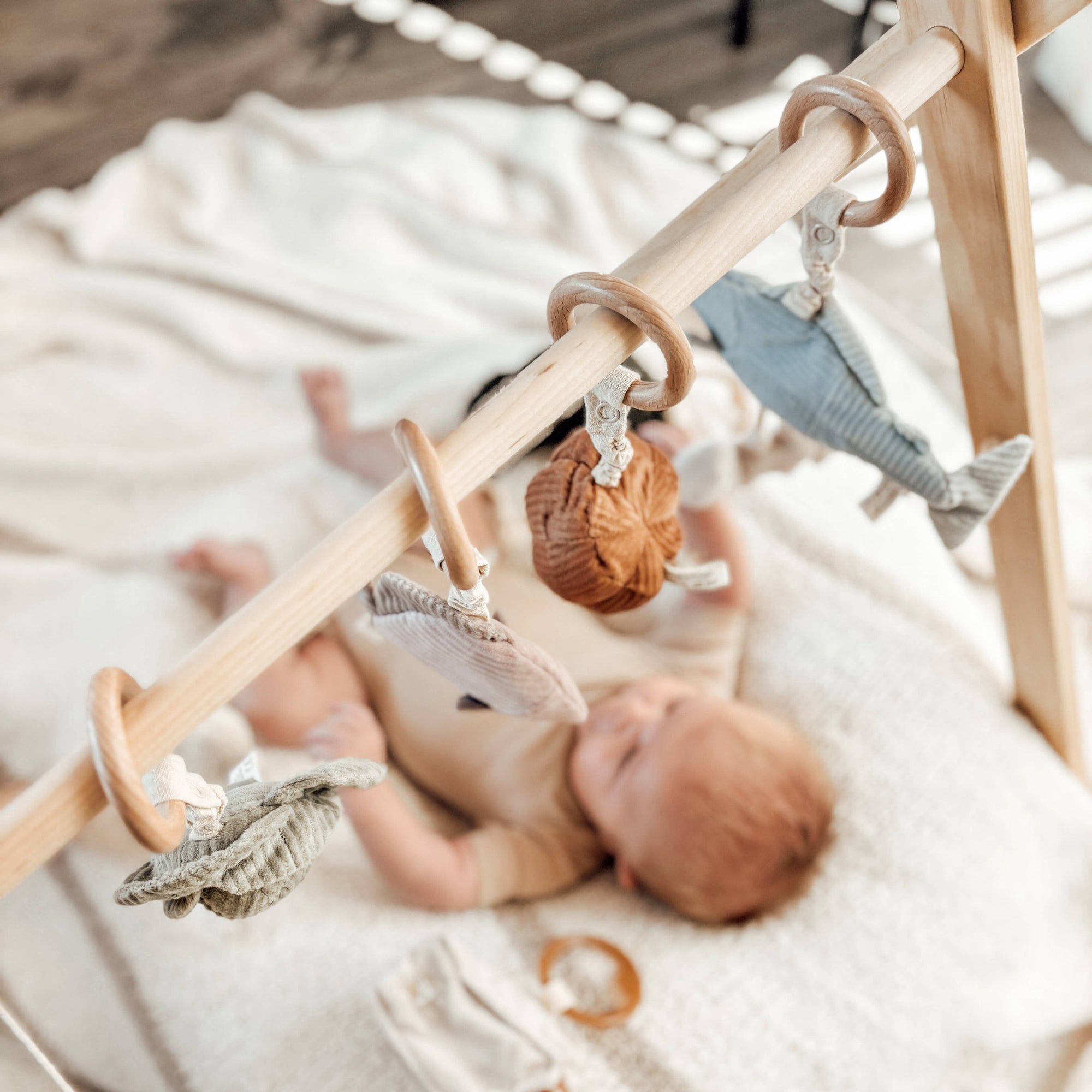 Baby lying on a white blanket with a wooden crib toy bar featuring hanging toys.