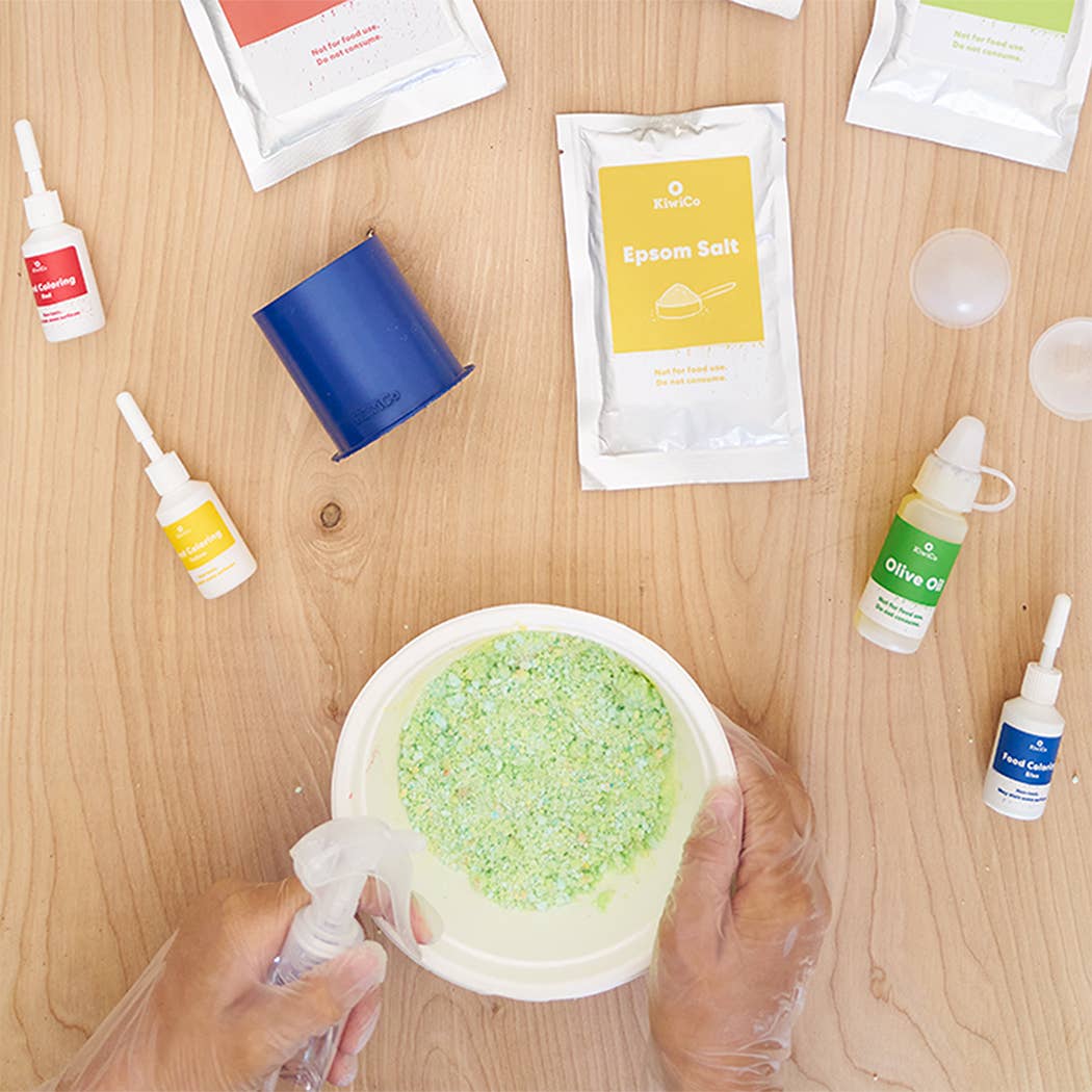 Person applying a green substance from a container with various skincare products on a wooden surface.