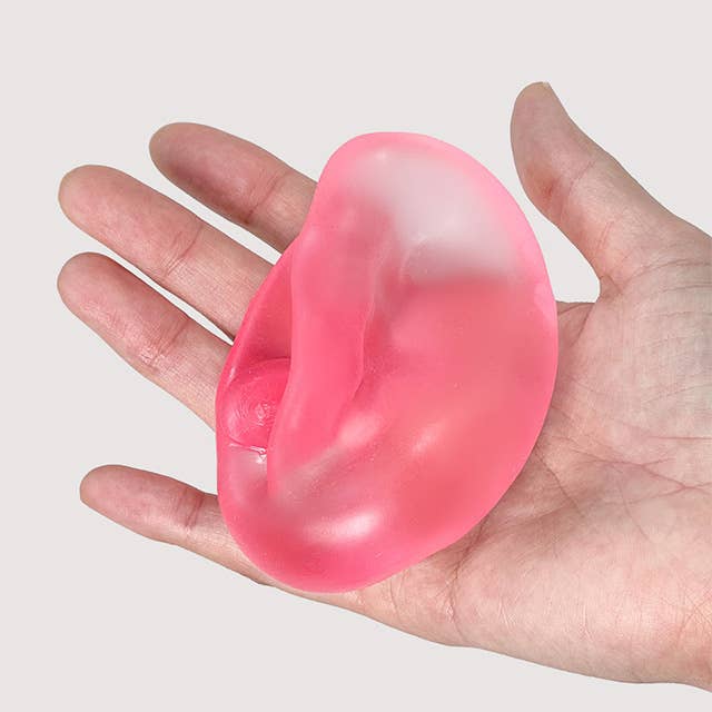 A translucent pink silicone ear-shaped object being held in a person's hand against a plain light gray background.