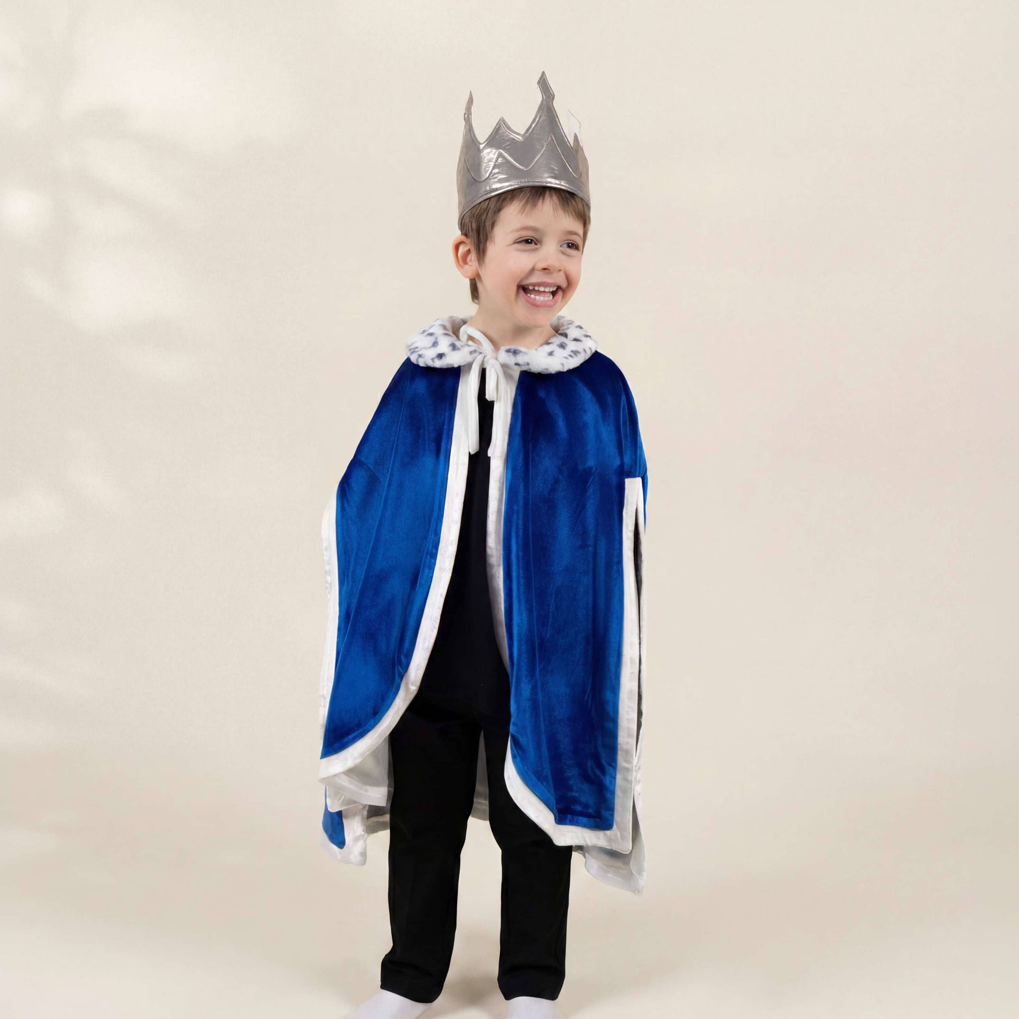 A young boy modeling a children's royal costume set, featuring a royal blue velvet cape with white trim, a matching silver crown, and a white patterned collar, paired with black pants and white socks, against a warm beige studio background.