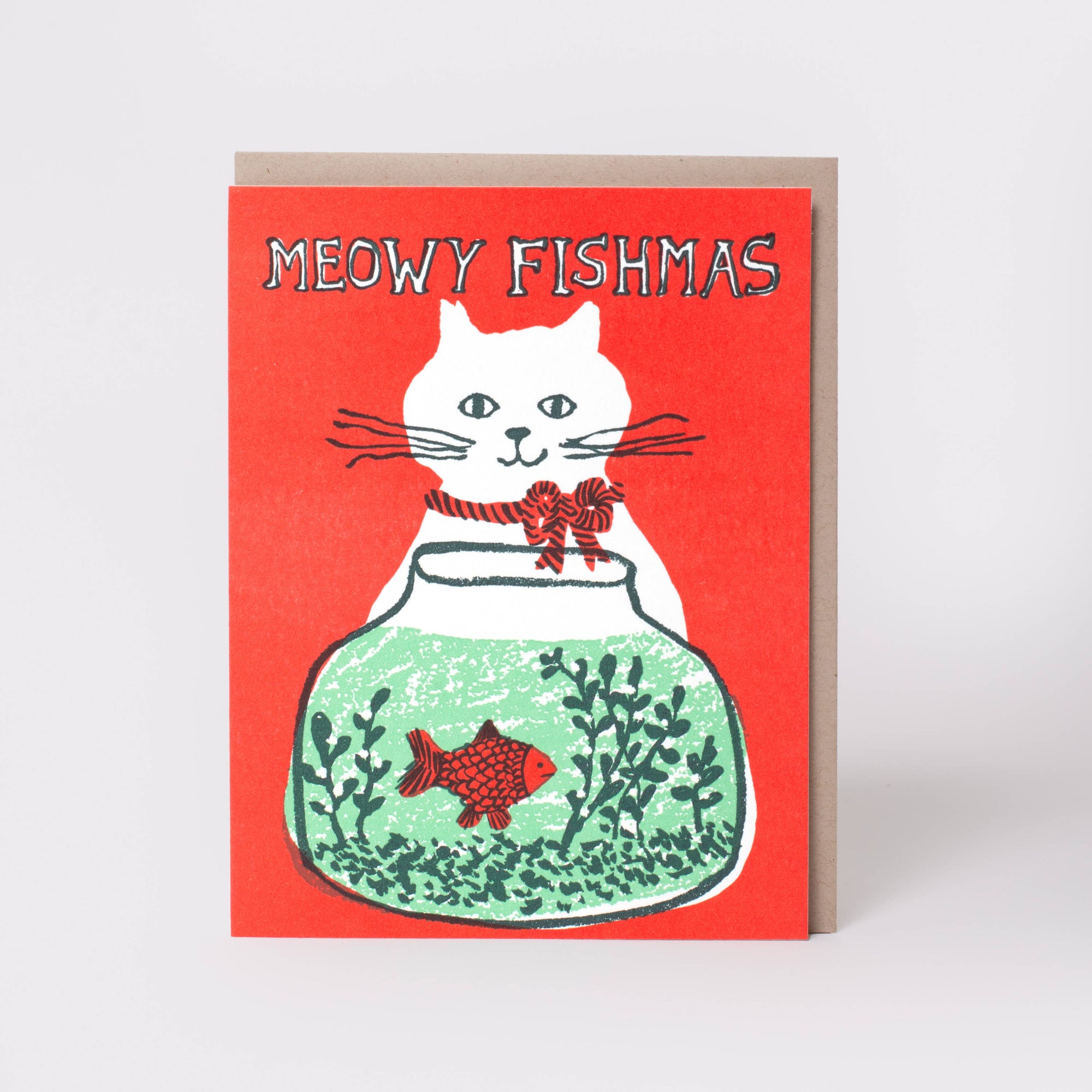 A greeting card with a red cover featuring a white cat sitting in a green fishbowl with a red fish, and the text 'MEOWY FISHMAS'. A brown envelope is visible behind the card on a light gray background.