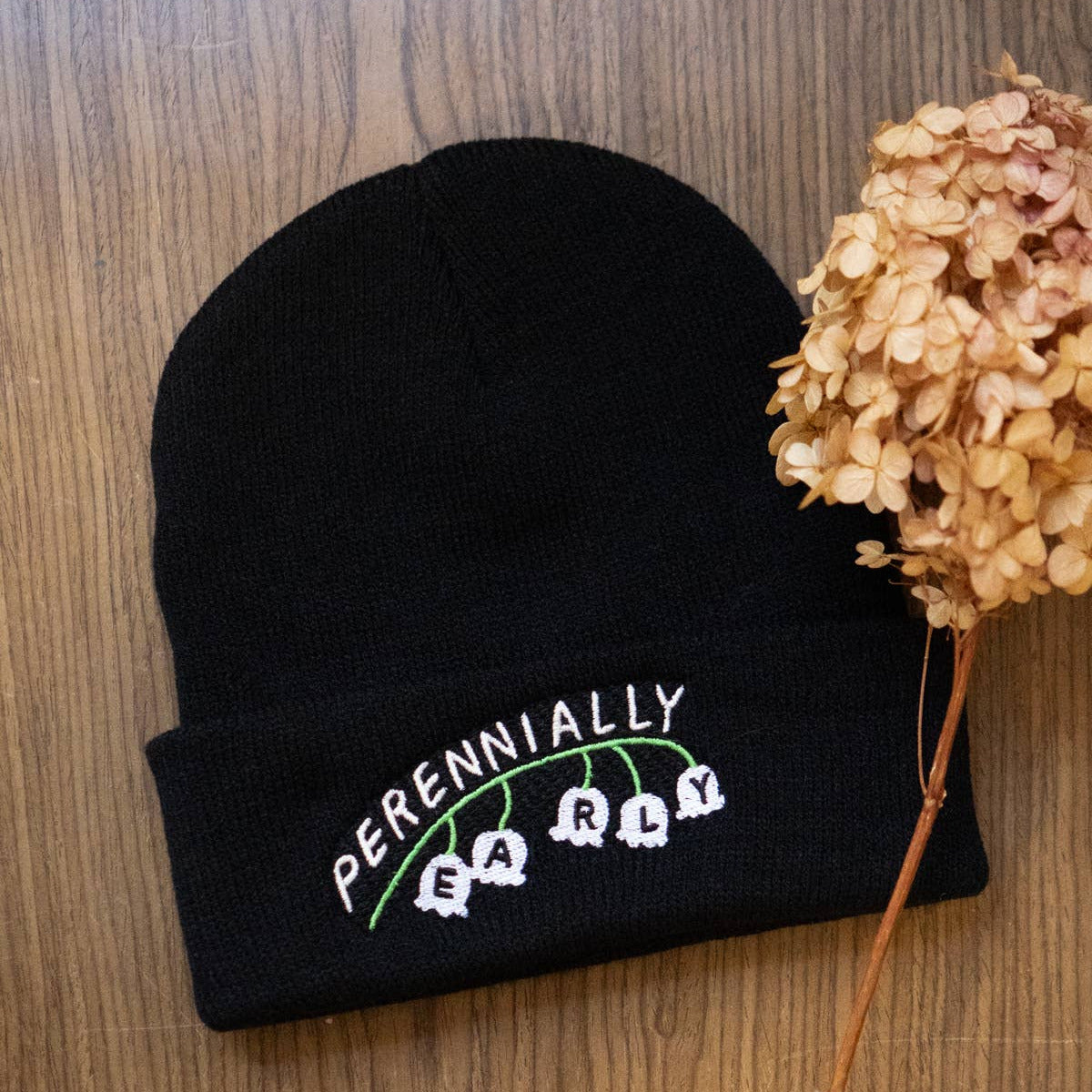 Black beanie with embroidered text on a wooden surface with a dried flower.