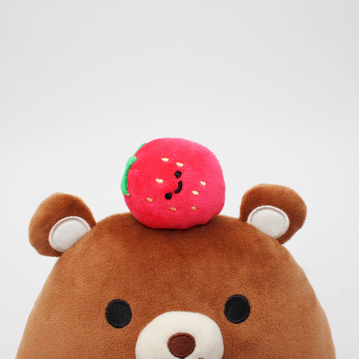 A close-up of a brown plush bear with a small pink strawberry toy resting on its head, photographed against a plain light gray background.