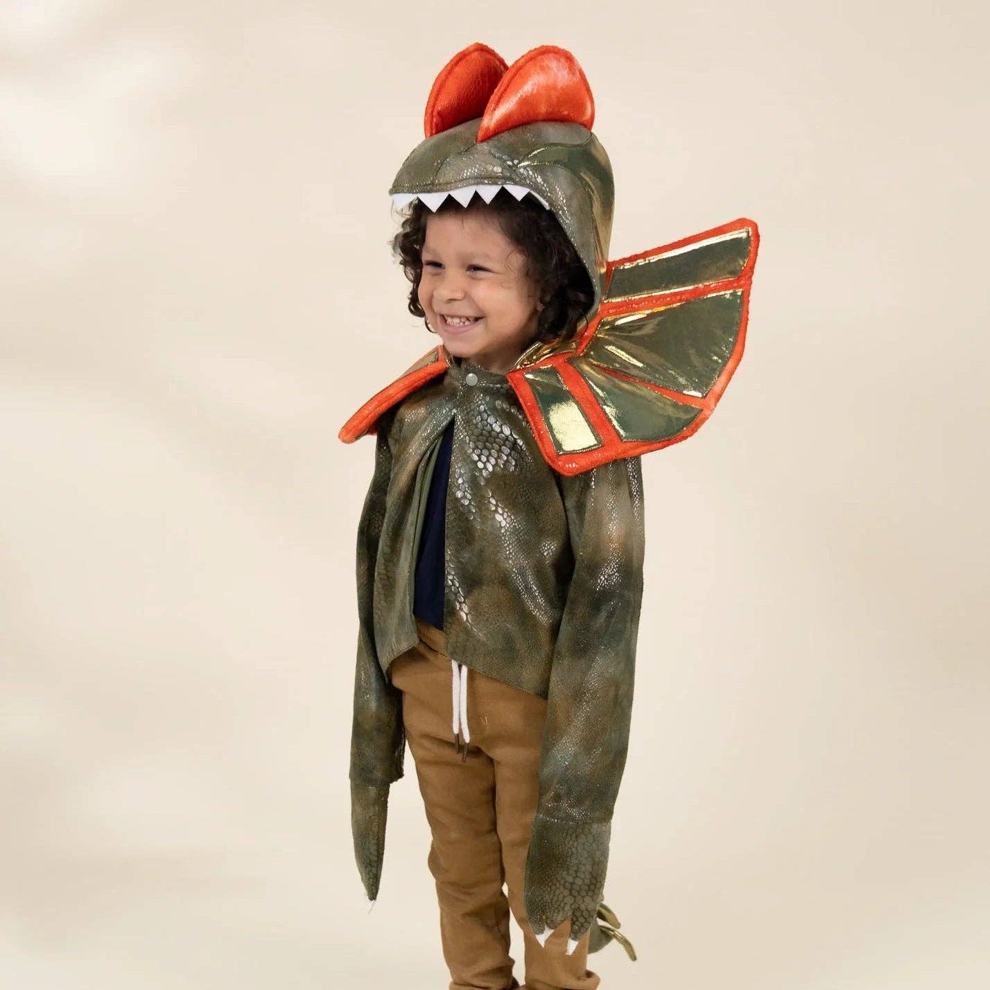 A child modeling a dinosaur costume consisting of a textured olive green hood with red horns and a white serrated mouth, a matching green cape with red trim, brown pants, and brown shoes, against a plain beige studio background.