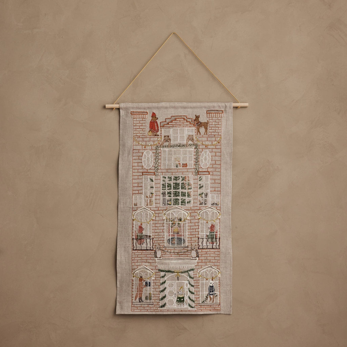 A vertical woven wall hanging tapestry featuring an embroidered-style facade of a brick building with windows, small figures, and animals, suspended from a wooden dowel against a textured beige wall.