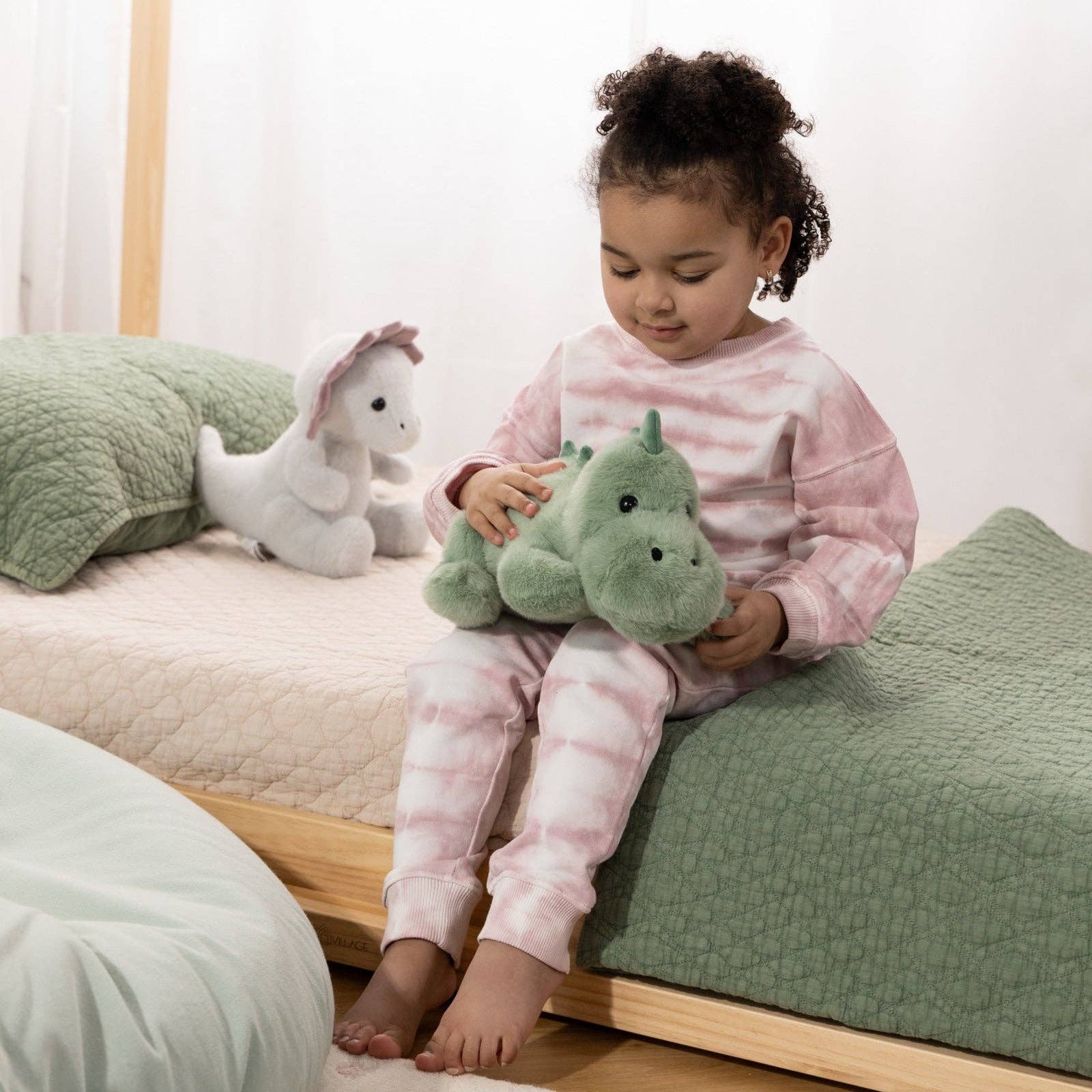 Child sitting on a bed holding a green plush toy, with another plush toy on the bed.