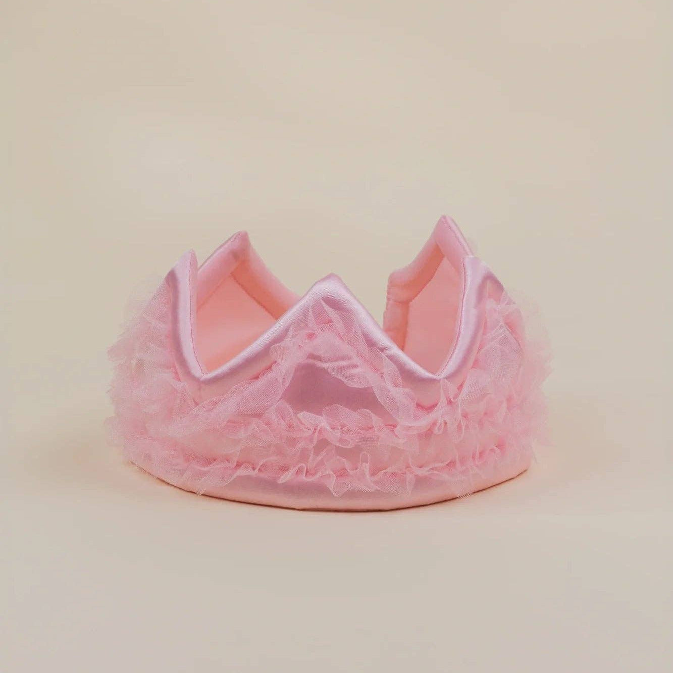 A pink ruffled tulle crown with a satin inner band, photographed on a plain beige studio background.