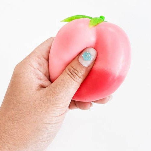 A hand squeezing a pink and red peach-shaped squishy toy with small green leaves on top, shown against a plain white background.