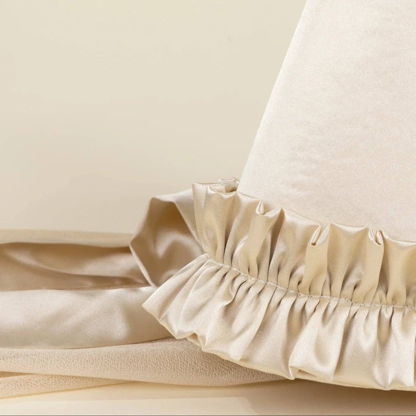 Close-up of a cream-colored fabric cone or hat-like textile with a gathered ruffled trim, resting on a matching satin fabric against a warm beige background.