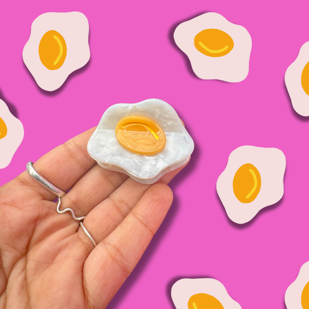 Hand holding a small fried egg-shaped object against a pink background with more egg shapes.