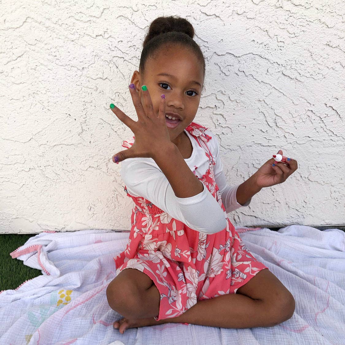 Young girl in a pink floral dress sitting on a blanket against a white wall.
