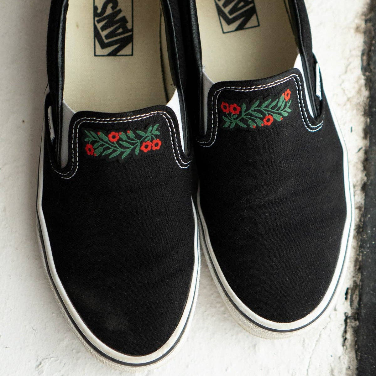 A pair of black Vans slip-on shoes with green and red floral embroidery on the side, displayed on a textured off-white surface.