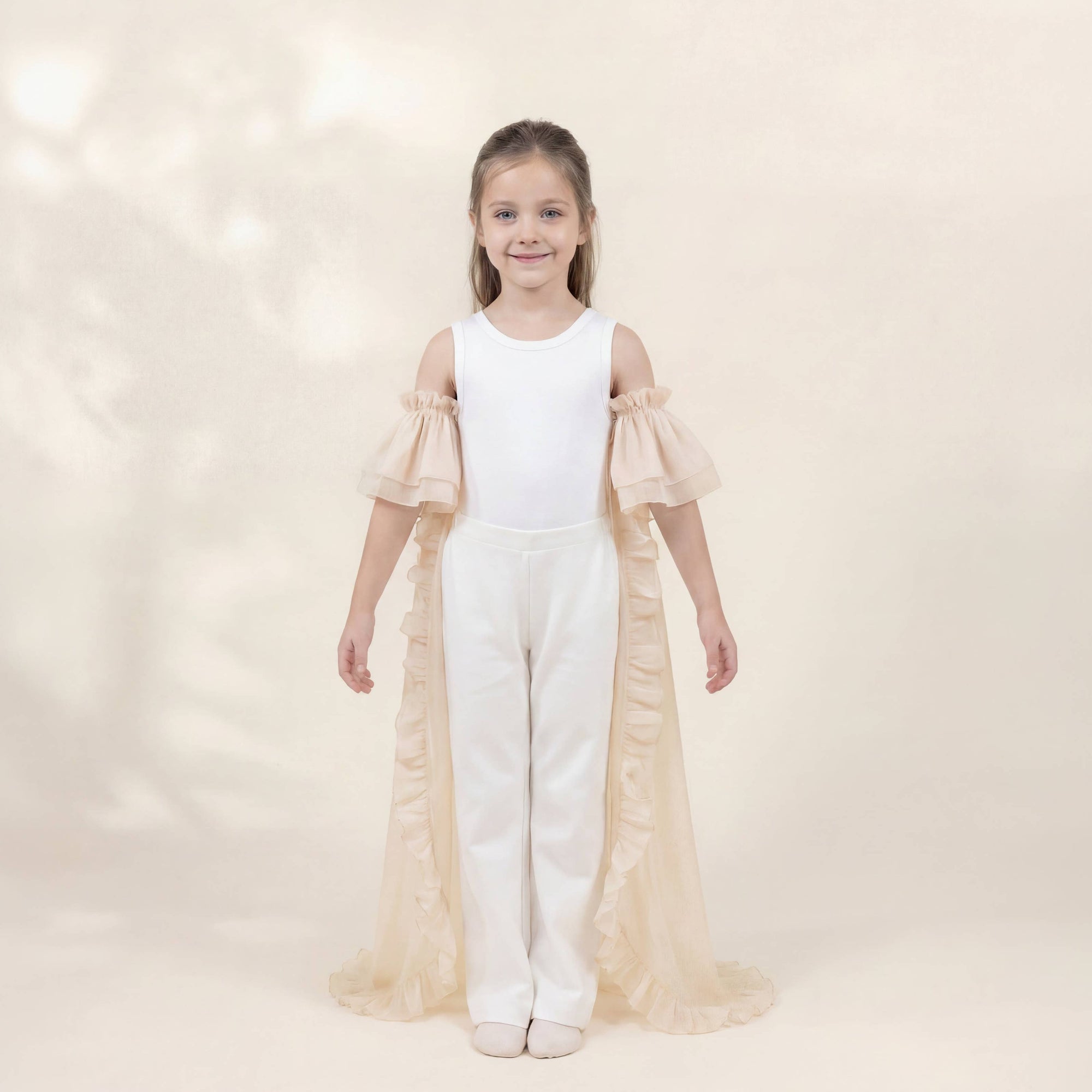 A young girl stands facing forward in a studio wearing a white sleeveless leotard with long ruffled sheer sleeves and white pants, layered with a long cream tulle cape. The background is a warm cream color with soft leafy shadows.