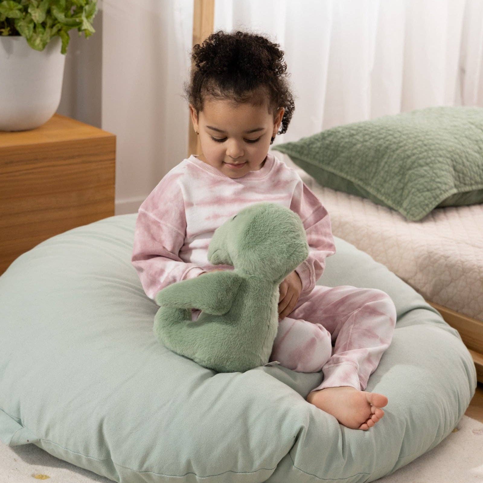 Child sitting on a green cushion holding a green plush toy in a room with white curtains and a plant.