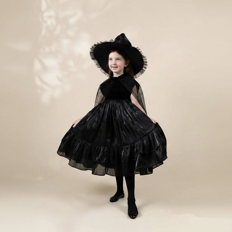 A young girl modeling a black witch costume consisting of a tiered tulle skirt dress, a matching wide-brimmed hat with ruffled trim, sheer cape sleeves, and black tights, set against a warm beige studio background.