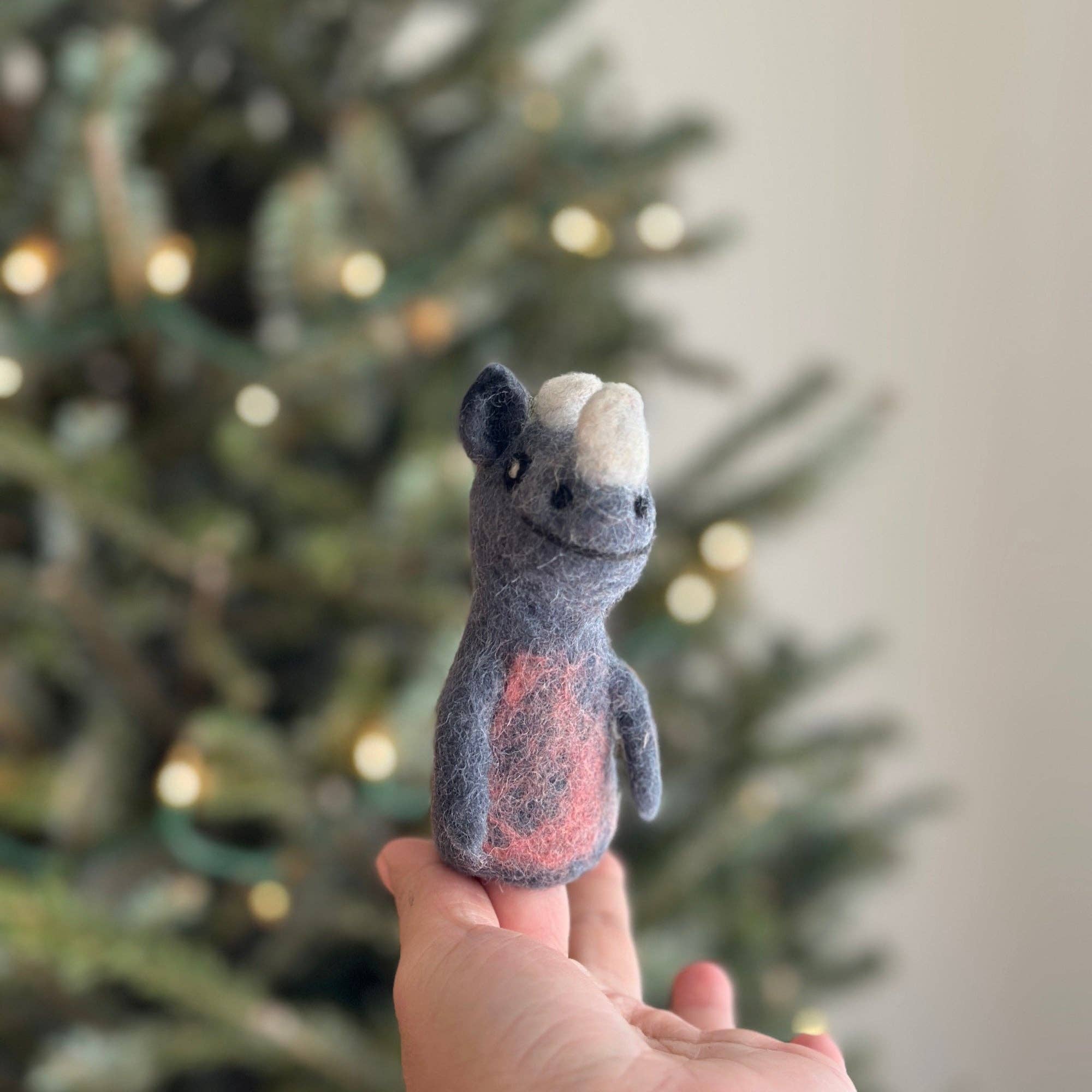 Hand holding a small felt rhino finger puppet in front of a decorated Christmas tree.