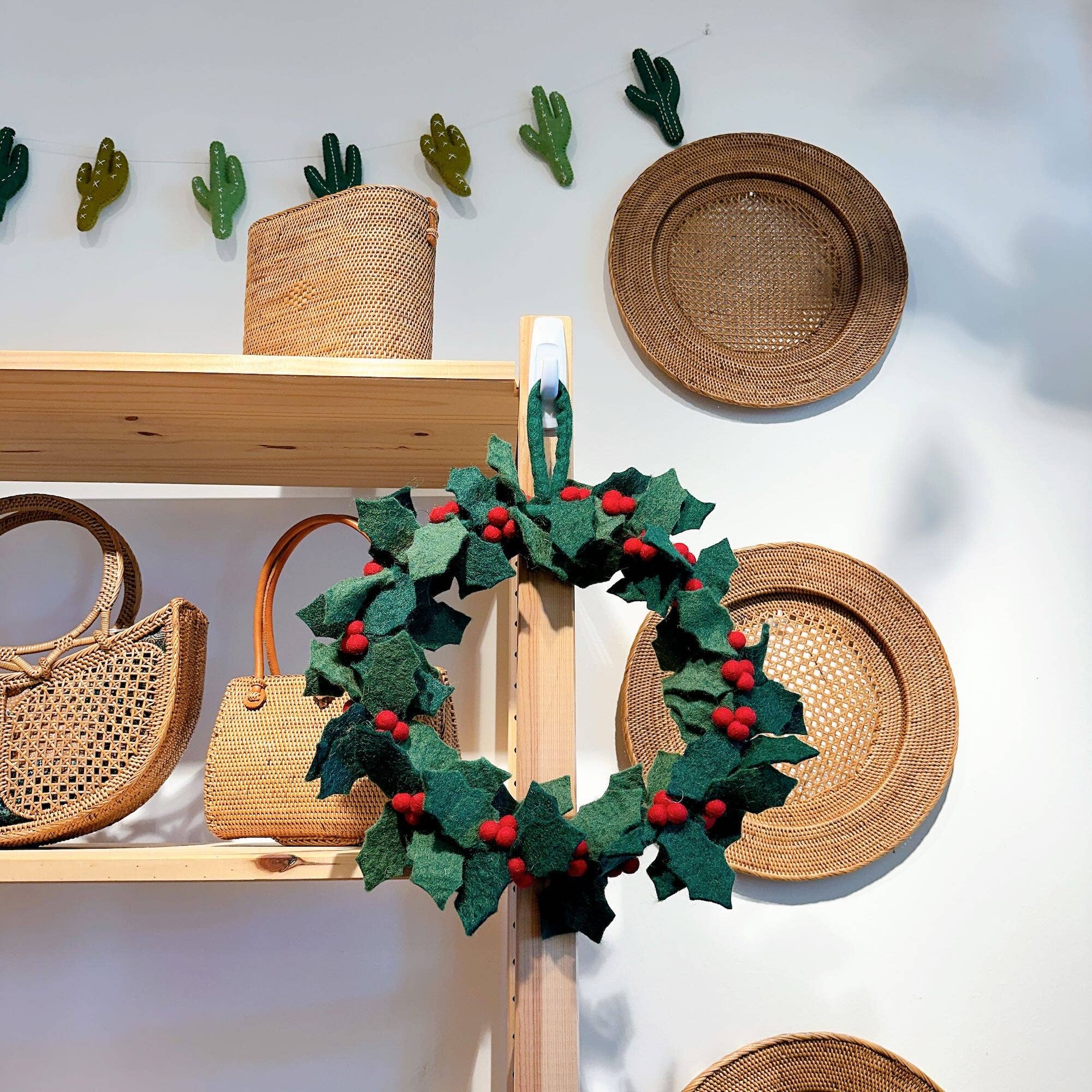 Felt holly wreath hanging on a wooden stand with wicker baskets and plates in the background.