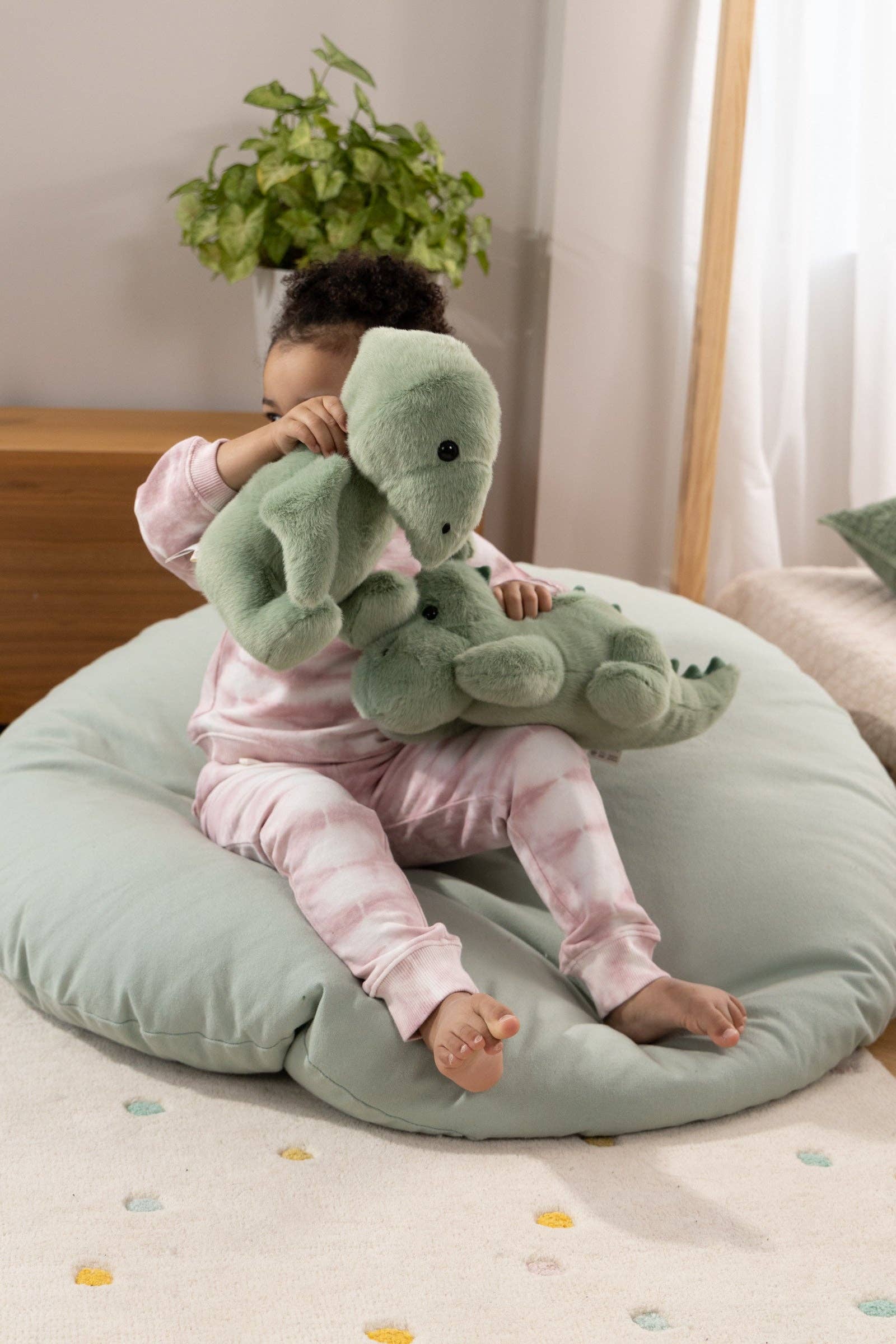 Child sitting on a green cushion holding a green plush toy in a cozy room.