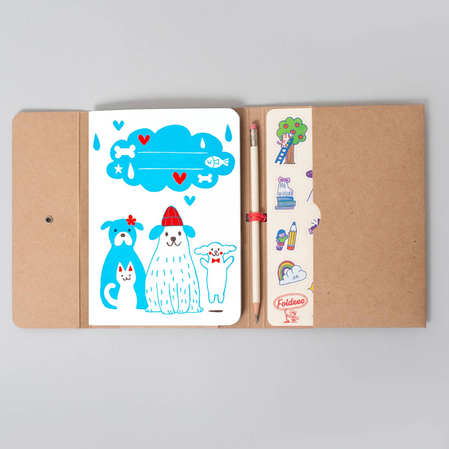 A kraft paper notebook or journal shown open on a light gray background, with a white insert page featuring cartoon dogs and a blue cloud illustration, plus a pencil and a vertical sticker strip on the right.
