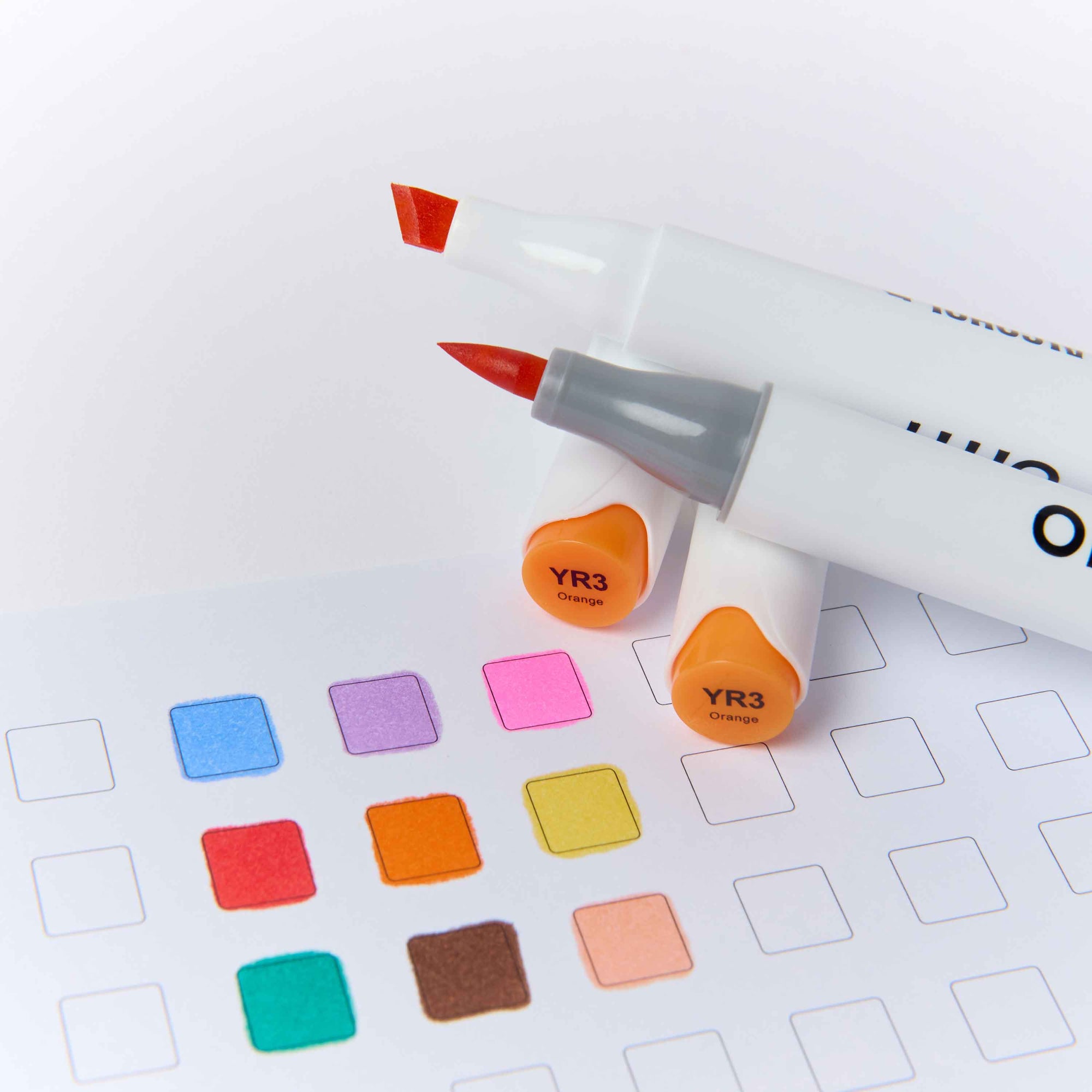 Two white dual-tip markers with orange caps and red tips, placed on top of a sheet of white paper with colored color swatches.