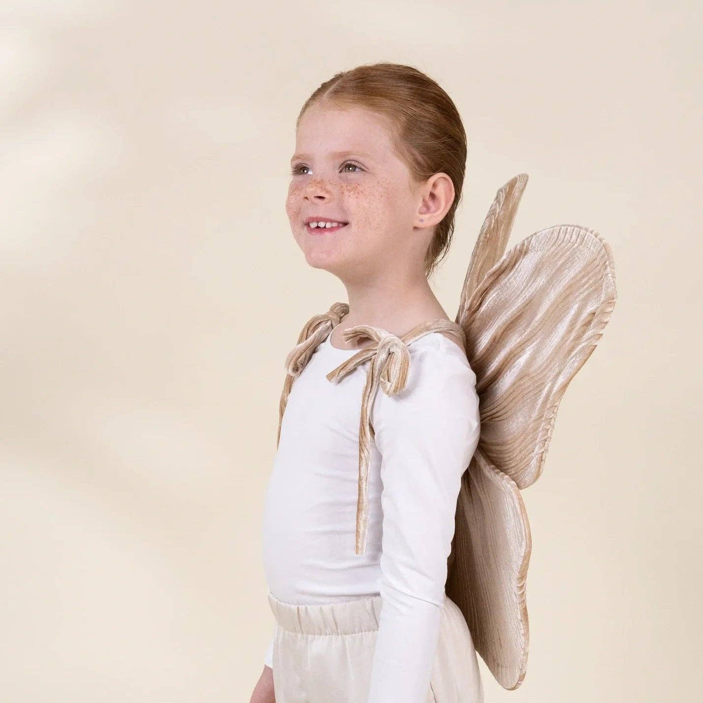 A young girl with light brown hair tied back, wearing a white long-sleeve top, cream pants, and a beige angel costume with matching wings, standing against a solid cream background.