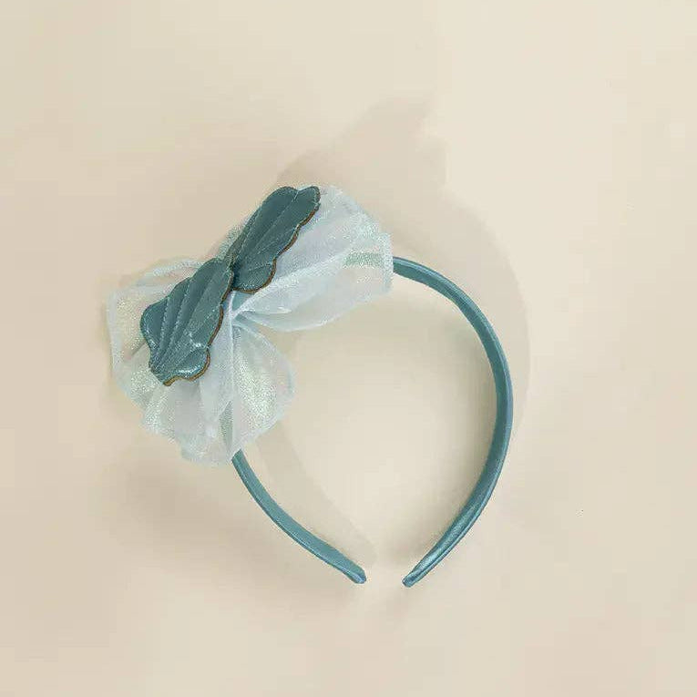 A teal blue plastic headband featuring a decorative floral embellishment on the left side with a dark teal ribbon bow and a sheer white tulle overlay, shown on a plain beige background.