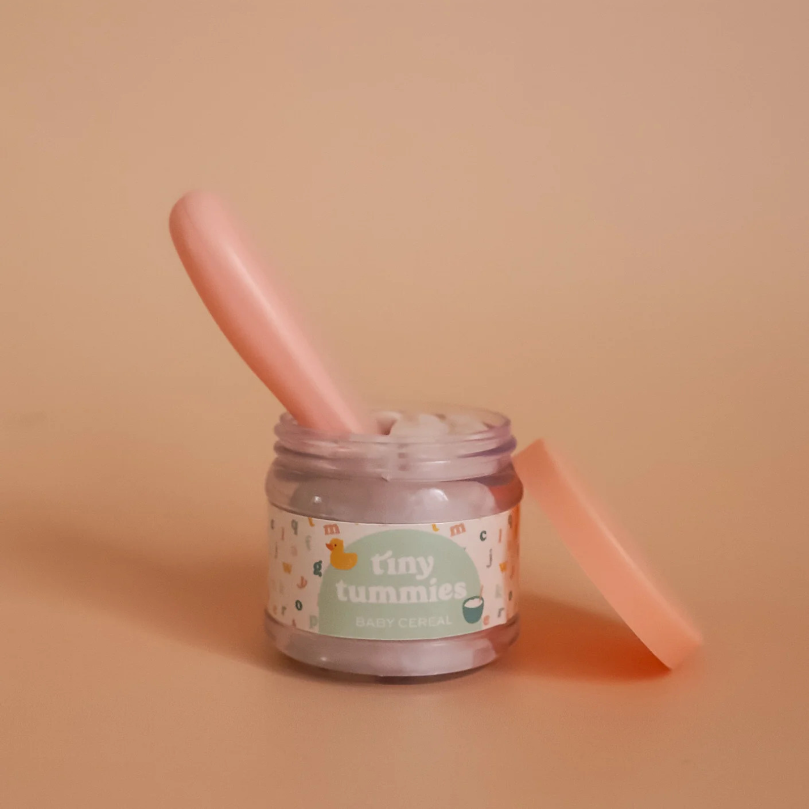 Jar of 'tiny tummies' baby food with a pink spoon on a beige background