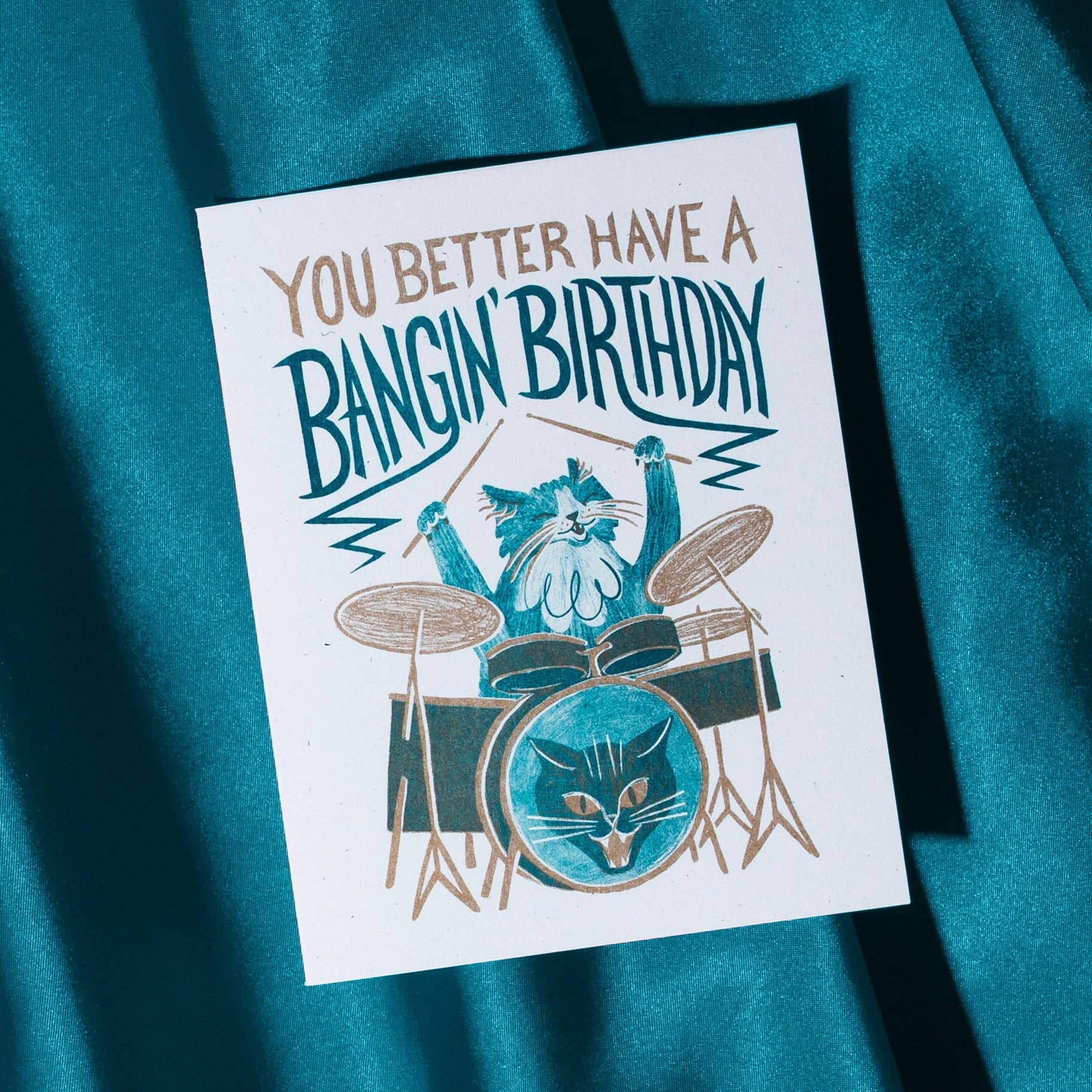 Greeting card with cat playing drums and text 'You better have a bangin' birthday' on a teal fabric background