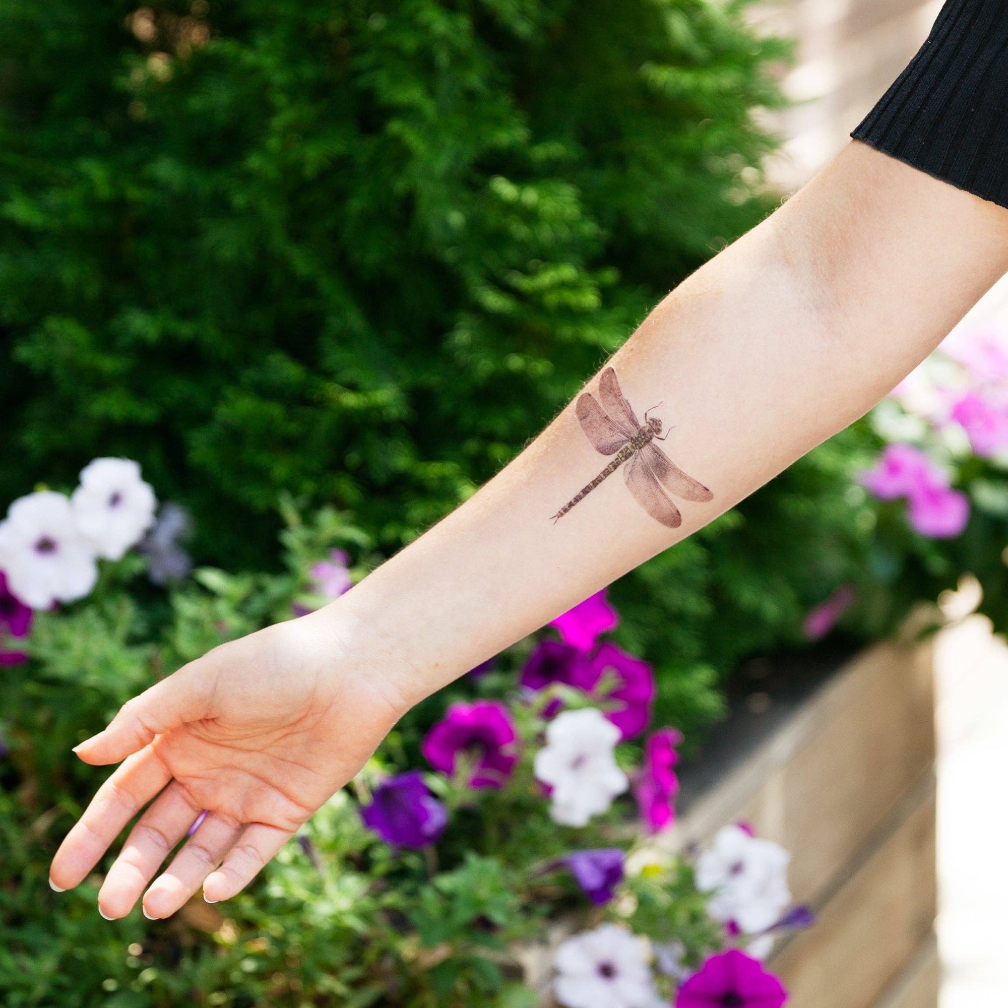 Person with a dragonfly tattoo on their arm, standing in front of flowers.
