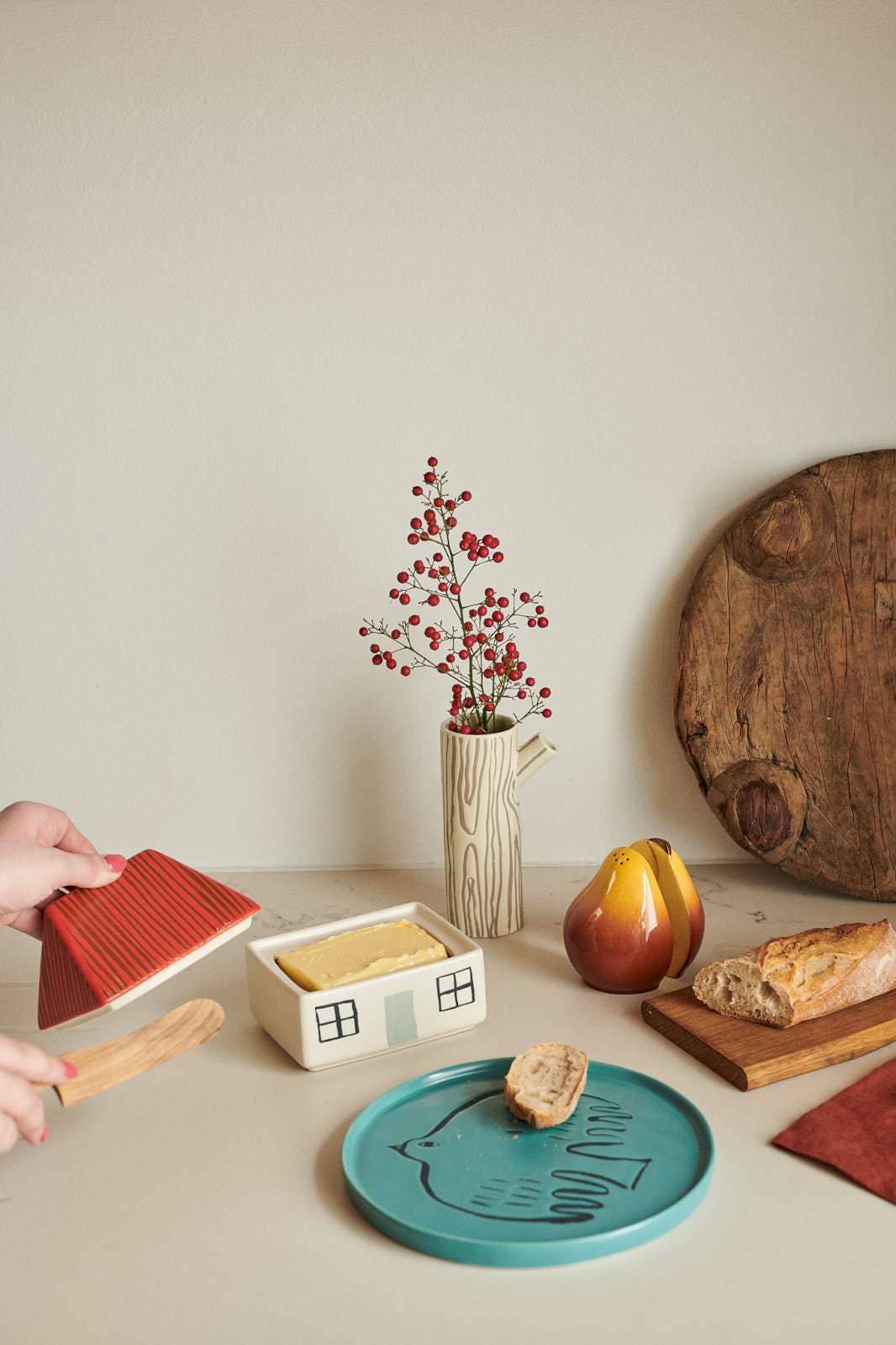A styled tabletop scene with a turquoise plate featuring a black cat silhouette, a small house-shaped dish with yellow wax melts, a red textured triangular dish, a wooden spoon, bread, a pear, and a vase with red berries against a warm beige background.