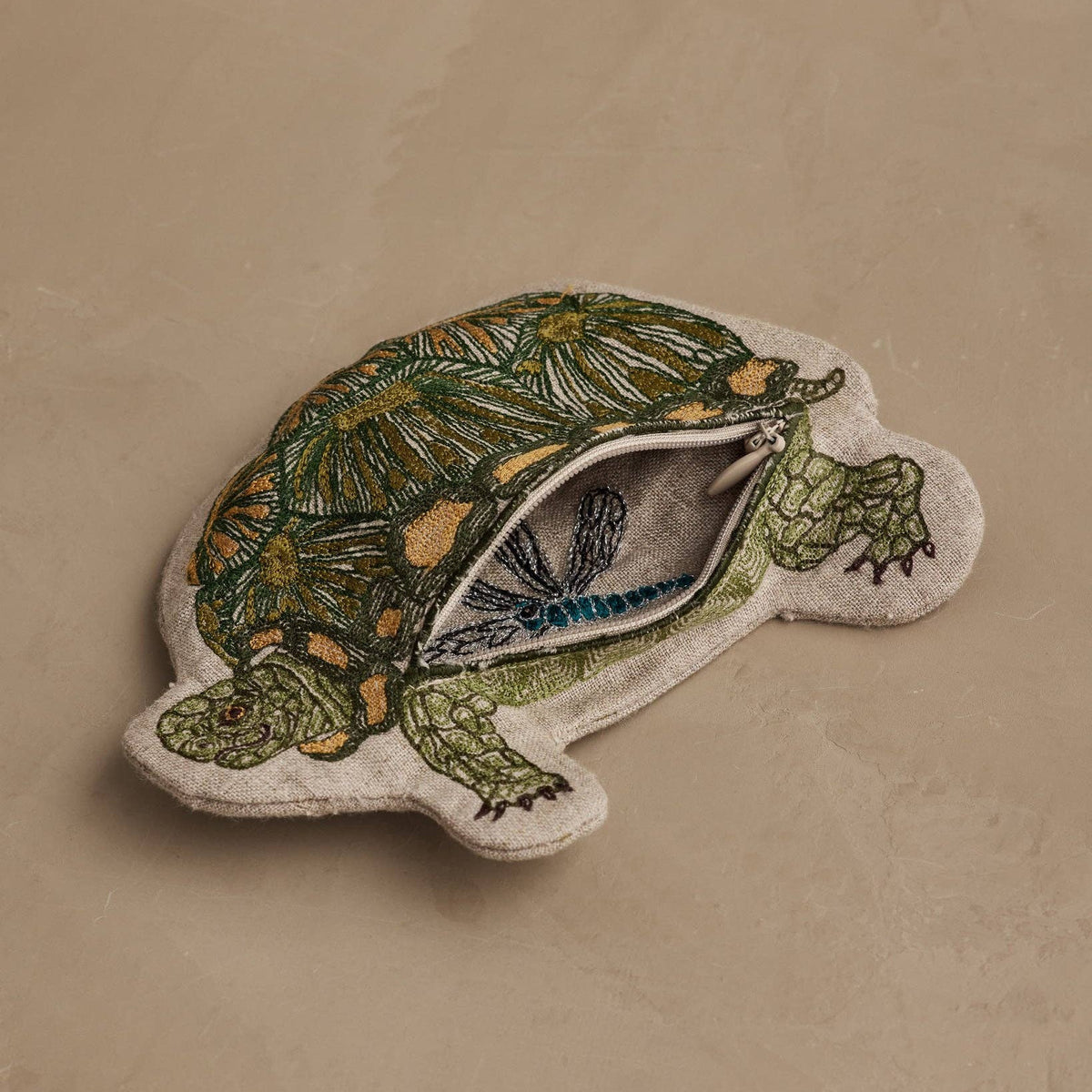 A turtle-shaped fabric pouch with a zipper, featuring an embroidered green and gold turtle design on a beige textured background.