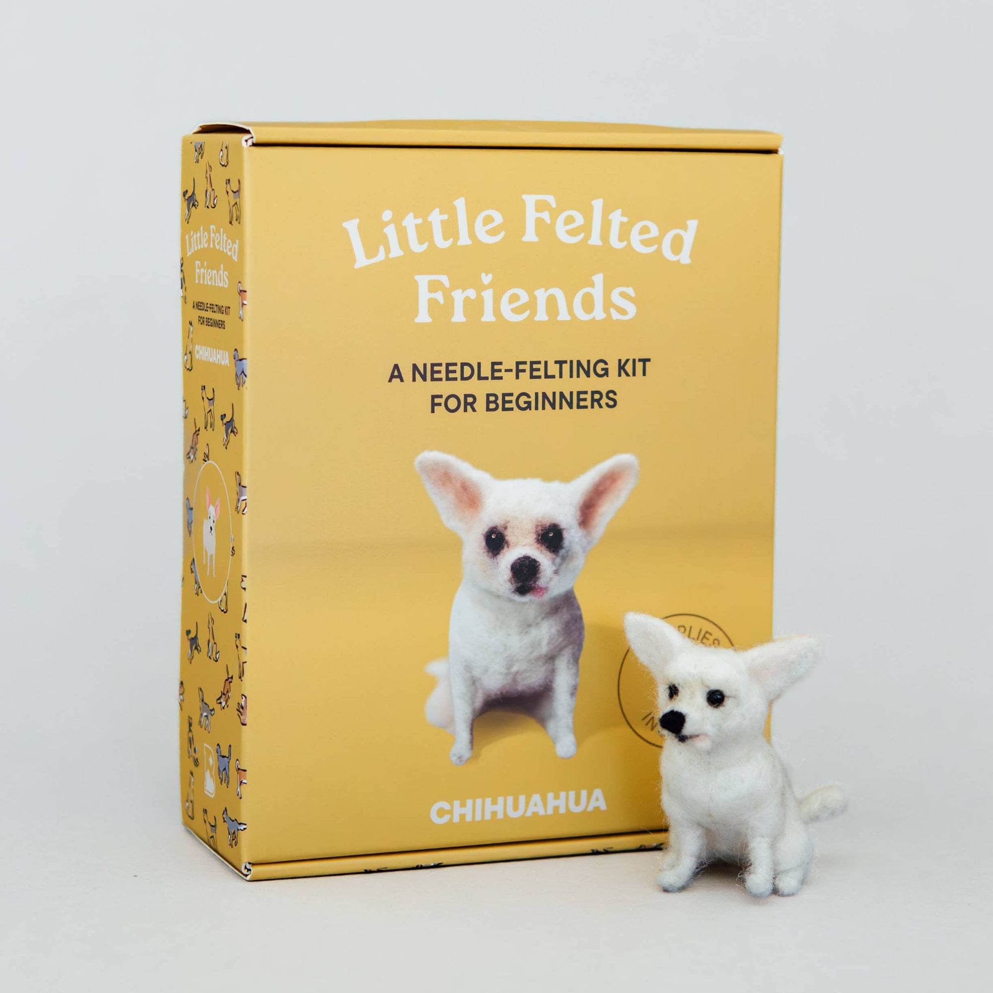 A needle felting kit box labeled 'Little Felted Friends' stands on a light gray studio background next to a small white chihuahua figurine. The box is mustard yellow with white text and a photo of a chihuahua on the front.