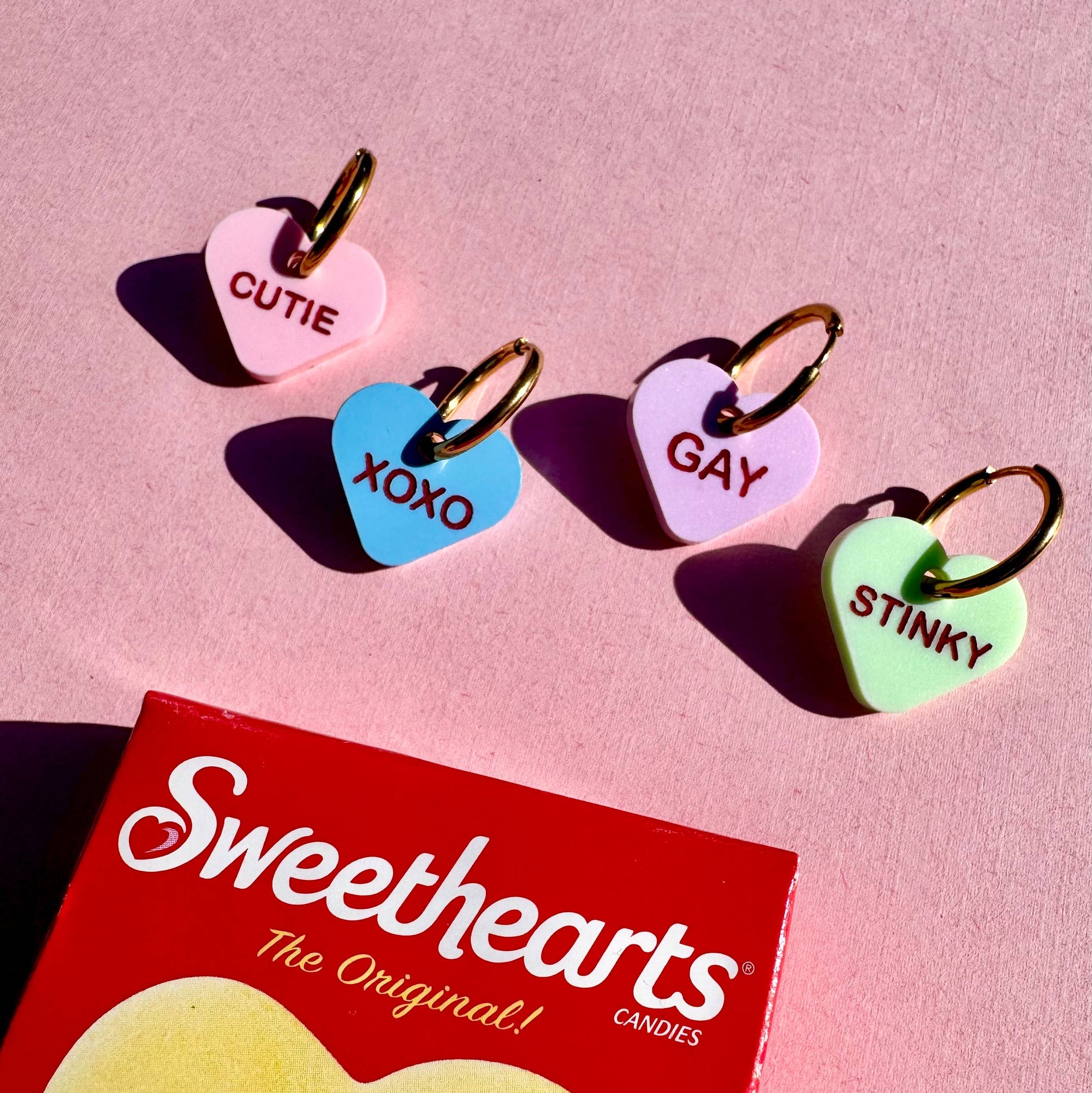 Heart-shaped candy charms with text on a pink background, featuring a box of Sweethearts candies.