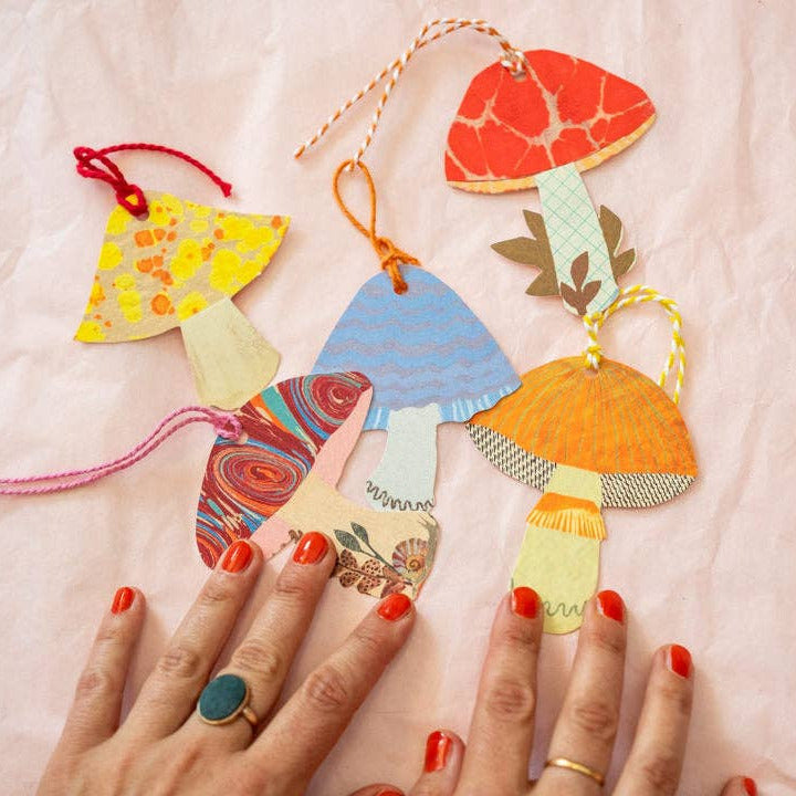 A set of colorful mushroom-shaped paper ornaments with hanging strings, arranged on a light pink crinkled paper background with two hands with red nail polish pointing at them.