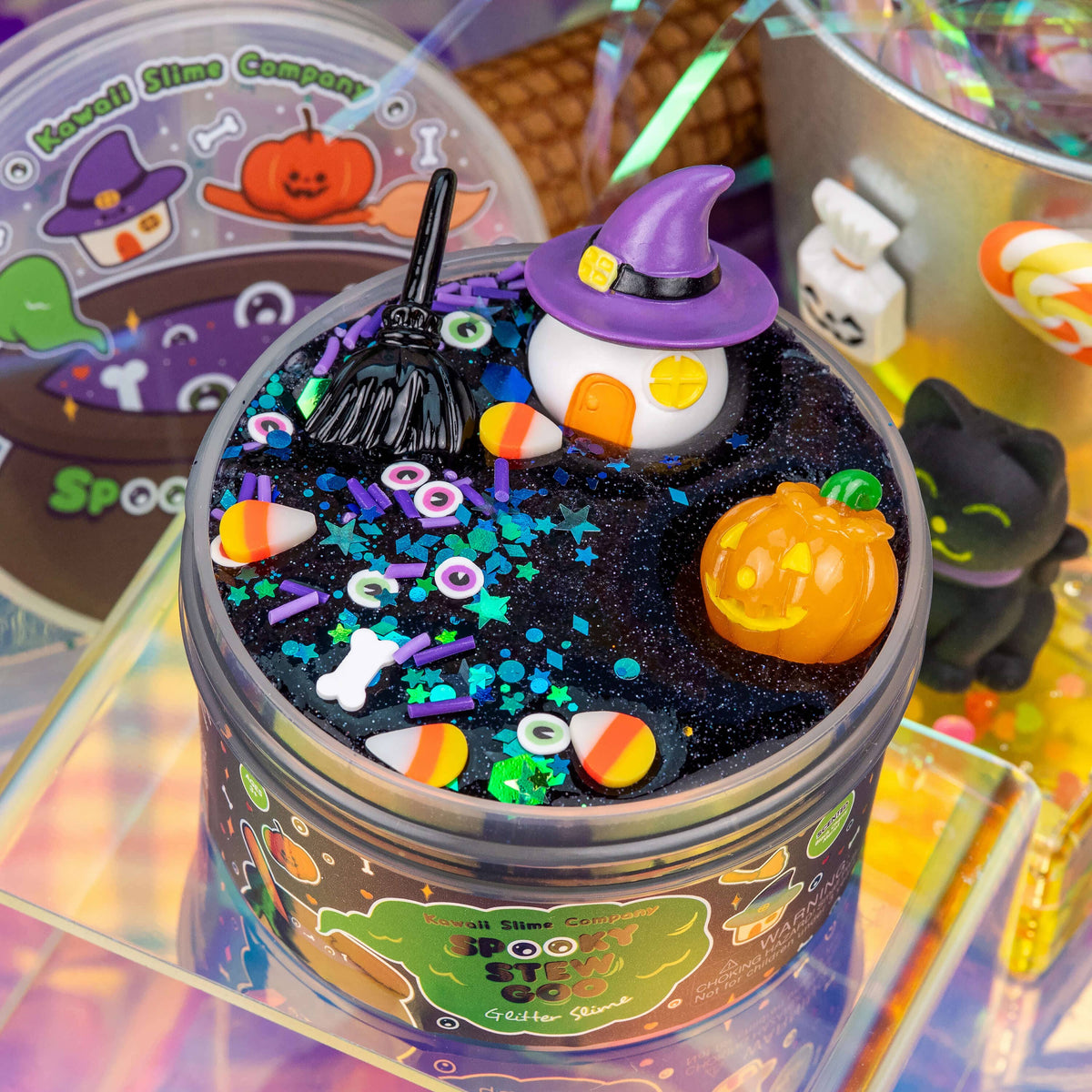 A round tin of Halloween-themed slime with a black glittery base, decorated with a small witch character, orange jack-o'-lantern, miniature broom, candy corn pieces, sprinkles, and colorful confetti. The tin label reads 'Spooky Slime Company' and 'Spooktacular'.