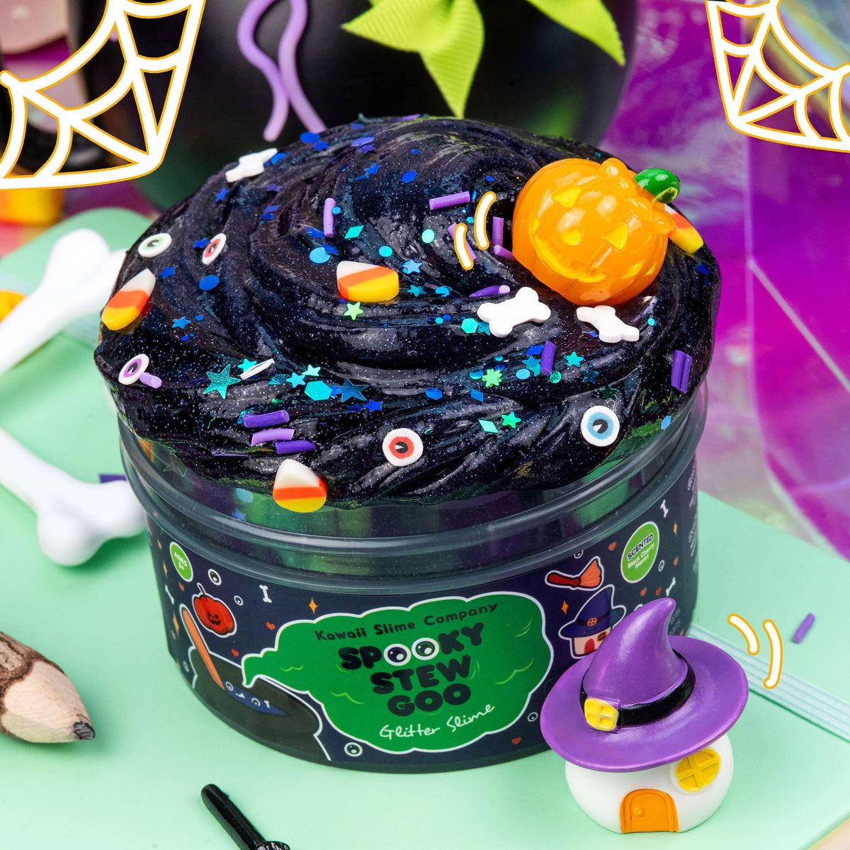 A Halloween-themed slime container filled with black glittery slime, decorated with colorful sprinkles, a small orange pumpkin, and candy corn pieces. The clear tub has a black label with green and orange 'Spooky Stew Good' branding, set on a mint green surface with decorative Halloween props including a small witch hat figure and neon spiderweb lights.