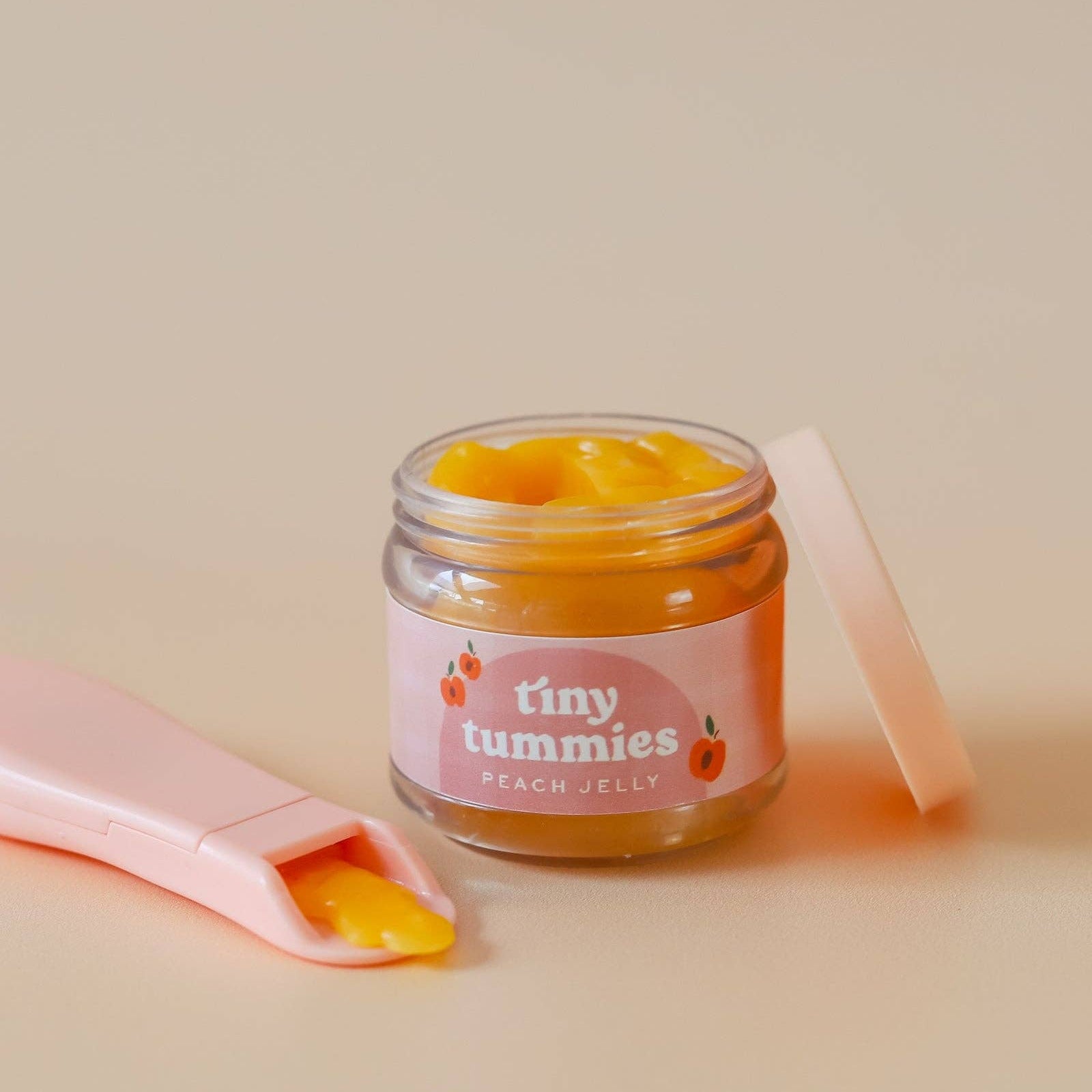 Jar of 'tiny tummies' peach jelly with a pink spoon on a beige background